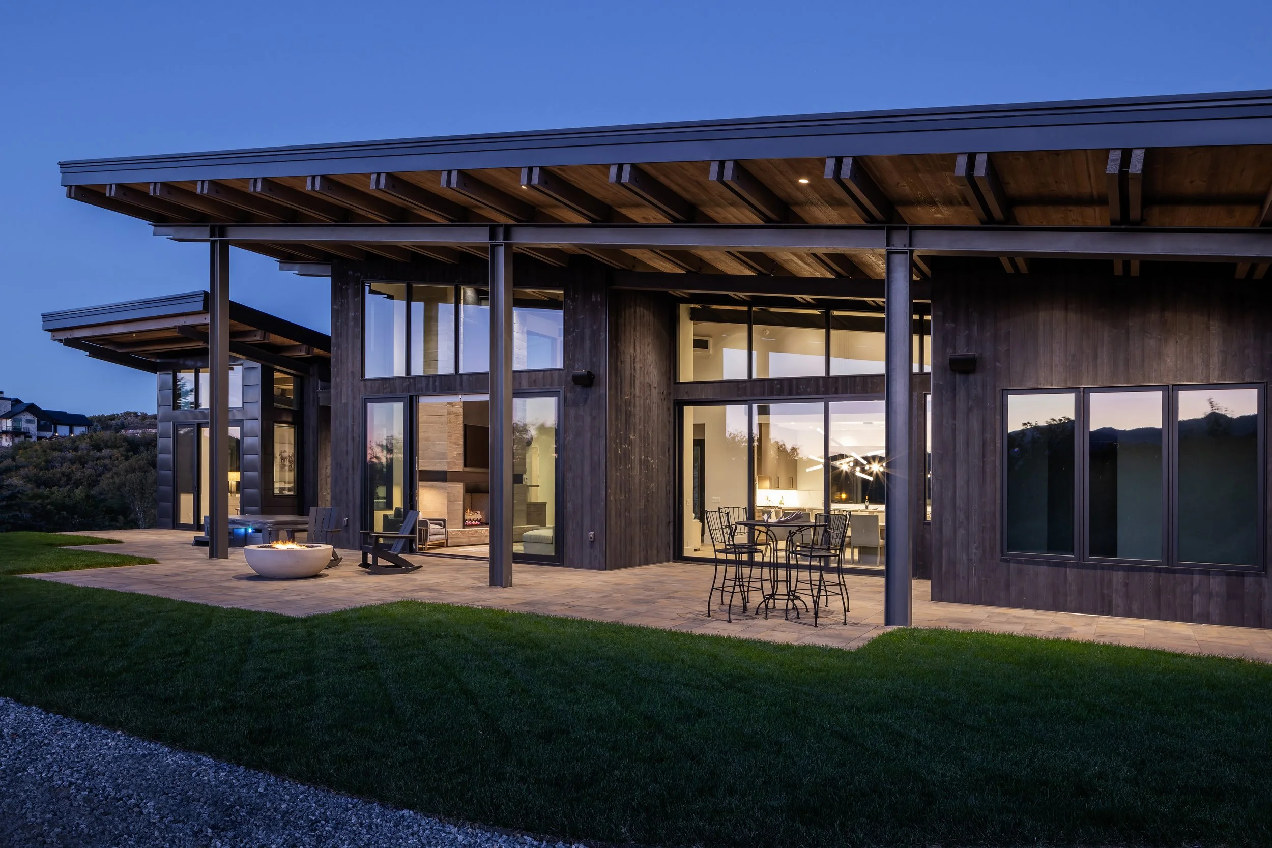 Modern house with large glass windows and a covered patio area, lit from inside, with outdoor seating and a fire pit on a grassy lawn during dusk.