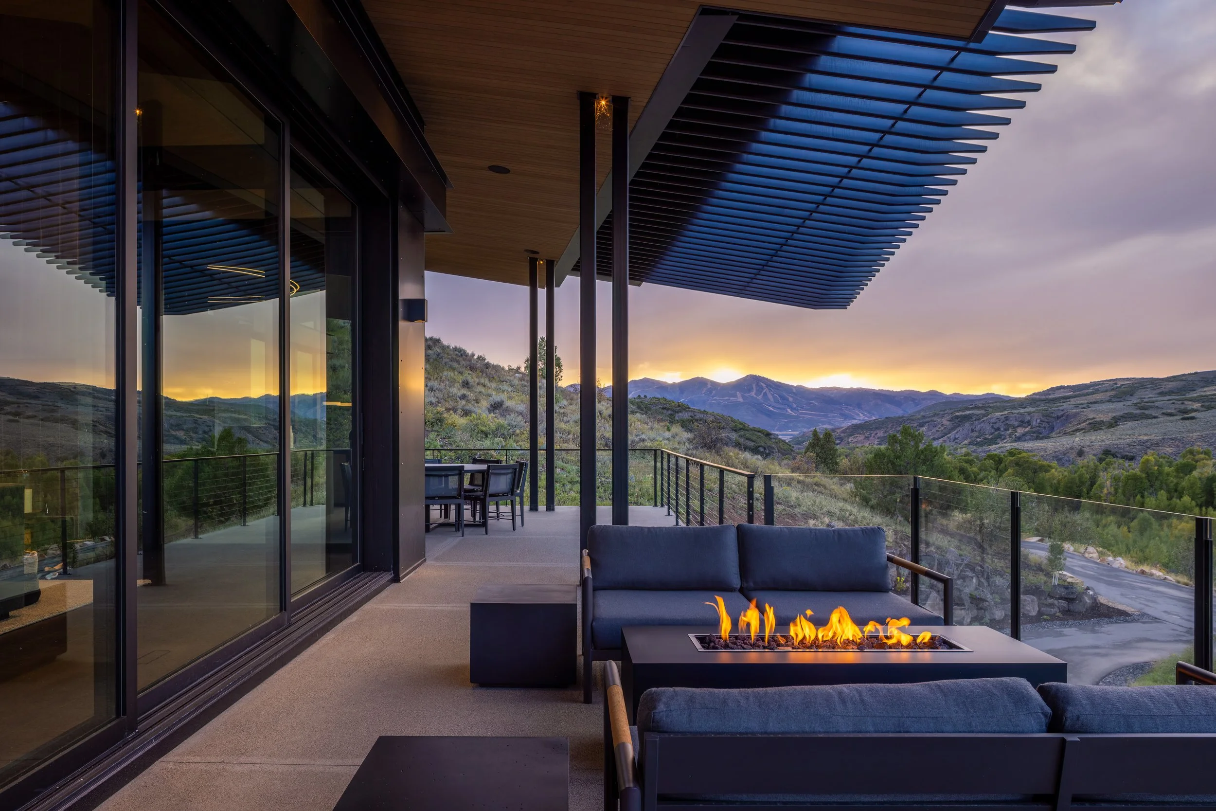A modern outdoor patio with glass sliding doors, a seating area with a fire pit, a table with chairs, and a mountain landscape in the background at sunset.