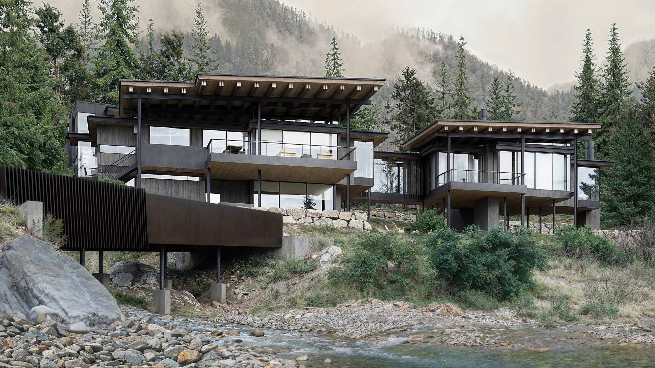 Modern house built on stilts with large glass windows, situated on a hillside surrounded by trees, overlooking a river with rocks and pebbles in the foreground.
