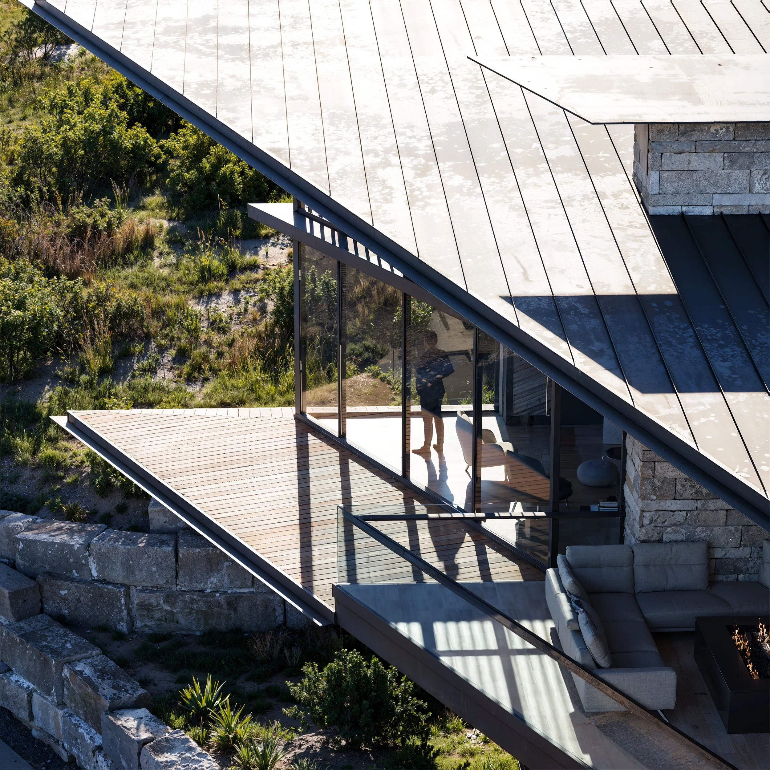 Modern house with large glass windows and metal roof, featuring a balcony and outdoor seating area, surrounded by lush greenery.