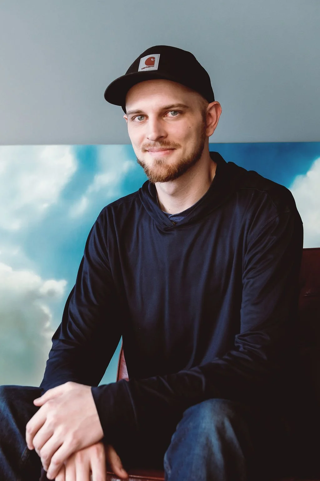 A young man with a beard and mustache wearing a black Carhartt baseball cap and a black hoodie, sitting indoors in front of a sky and cloud mural.