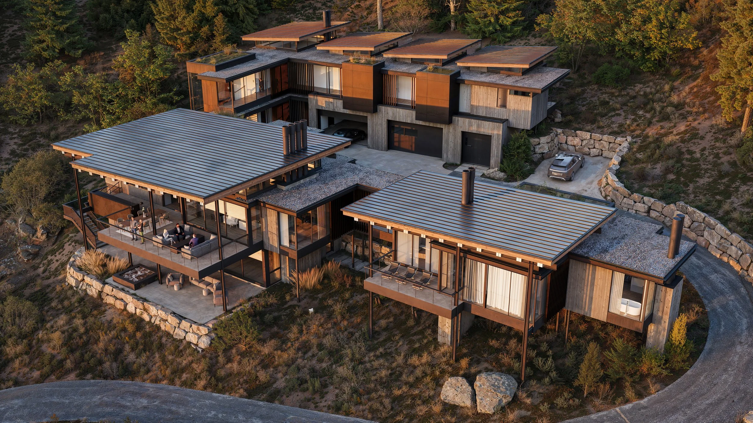 An aerial view of a modern multi-level house built on a hillside with large glass windows, wooden exterior, and metal roof sections. The house features outdoor patios with seating areas and a driveway with cars, surrounded by trees and rocky terrain.