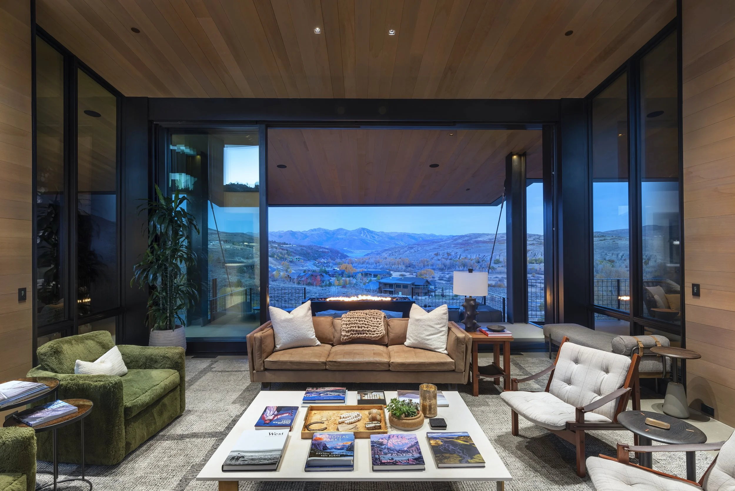 Modern living room with large windows showing mountain view, brown sofa with white and beige pillows, green armchair, white coffee table with books and decor, side table with lamp, and various seating arrangements.