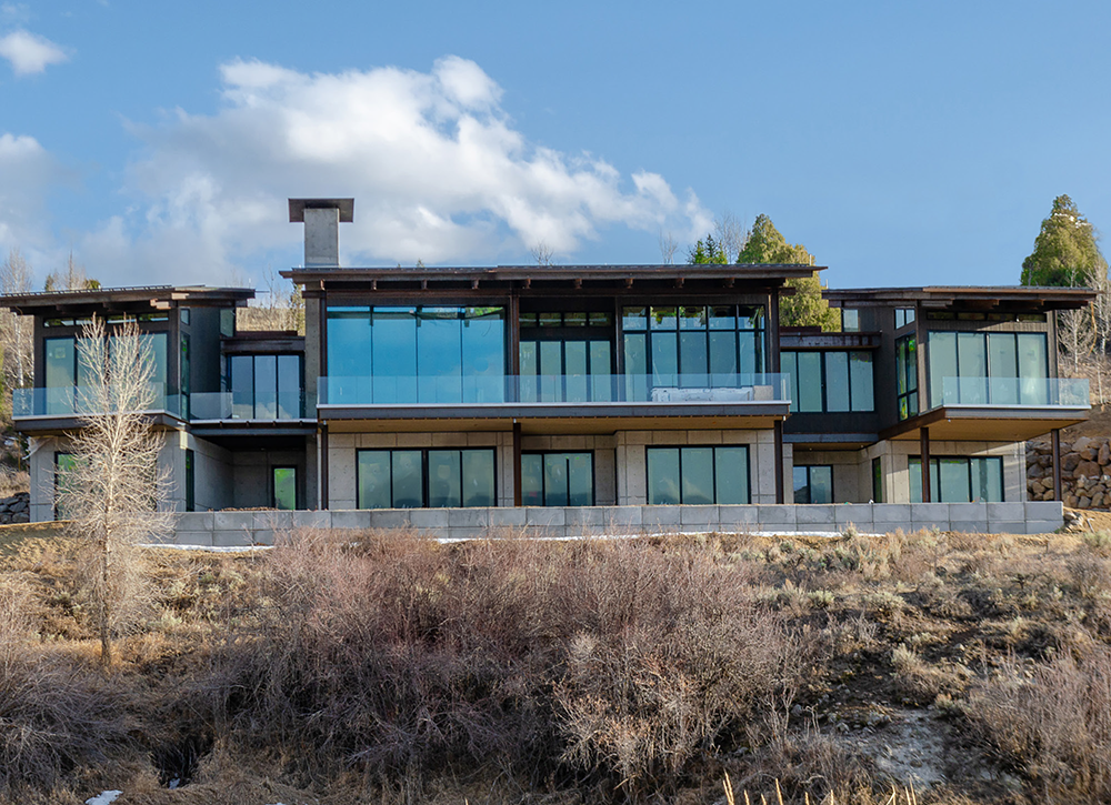 Modern house with large glass windows and balconies on a hillside under a partly cloudy sky.
