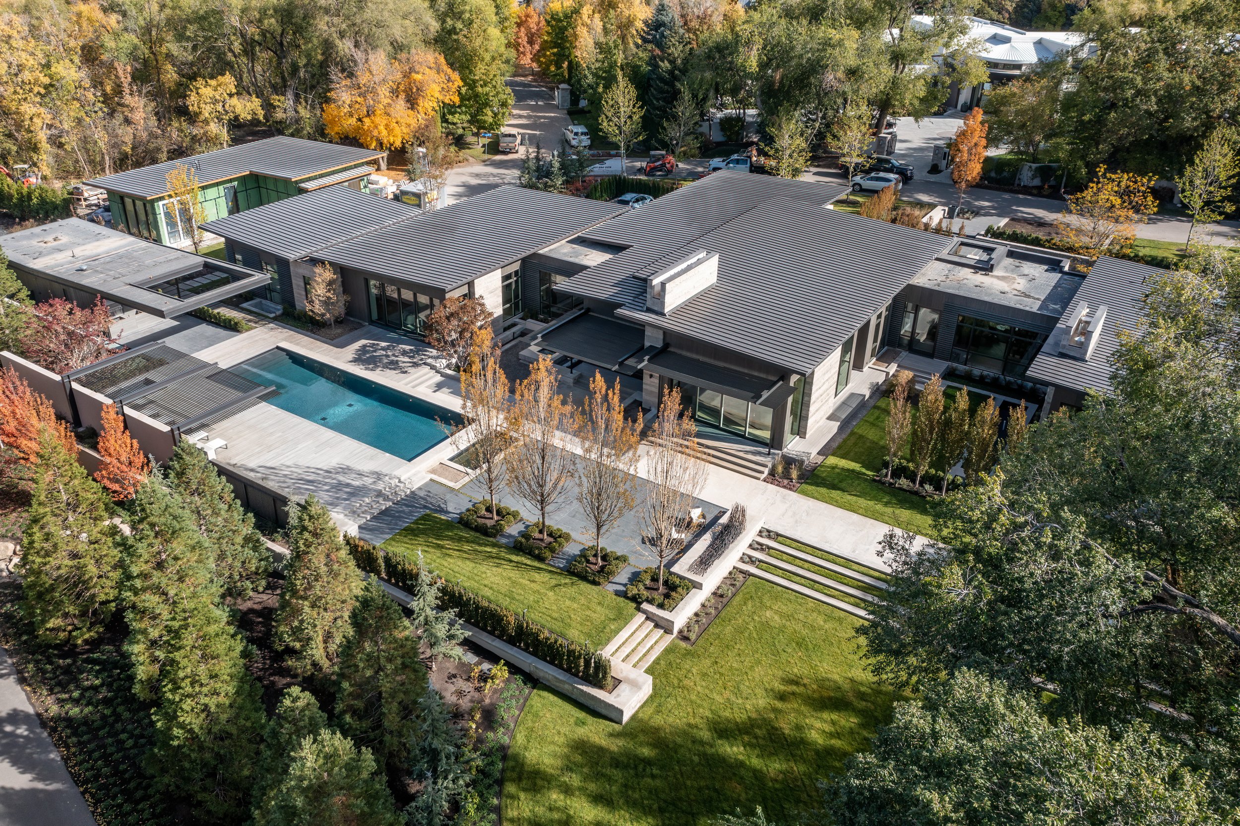A modern, large house with a flat roof, outdoor swimming pool, landscaped yard, and multiple outdoor seating areas, surrounded by trees with autumn foliage.