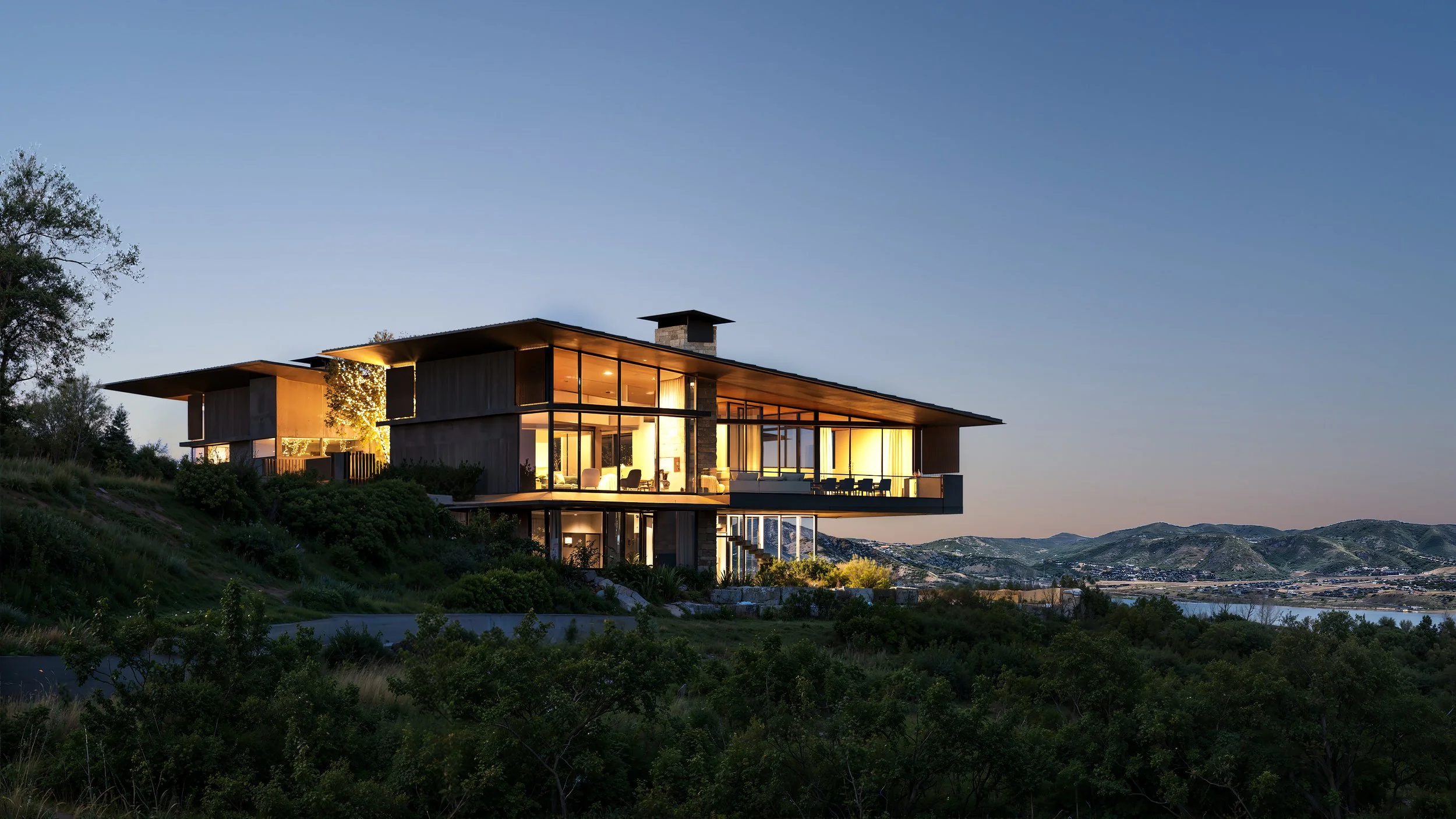 Modern house with large glass windows and a flat roof, lit from within, on a hillside during dusk with mountains and a body of water in the background.