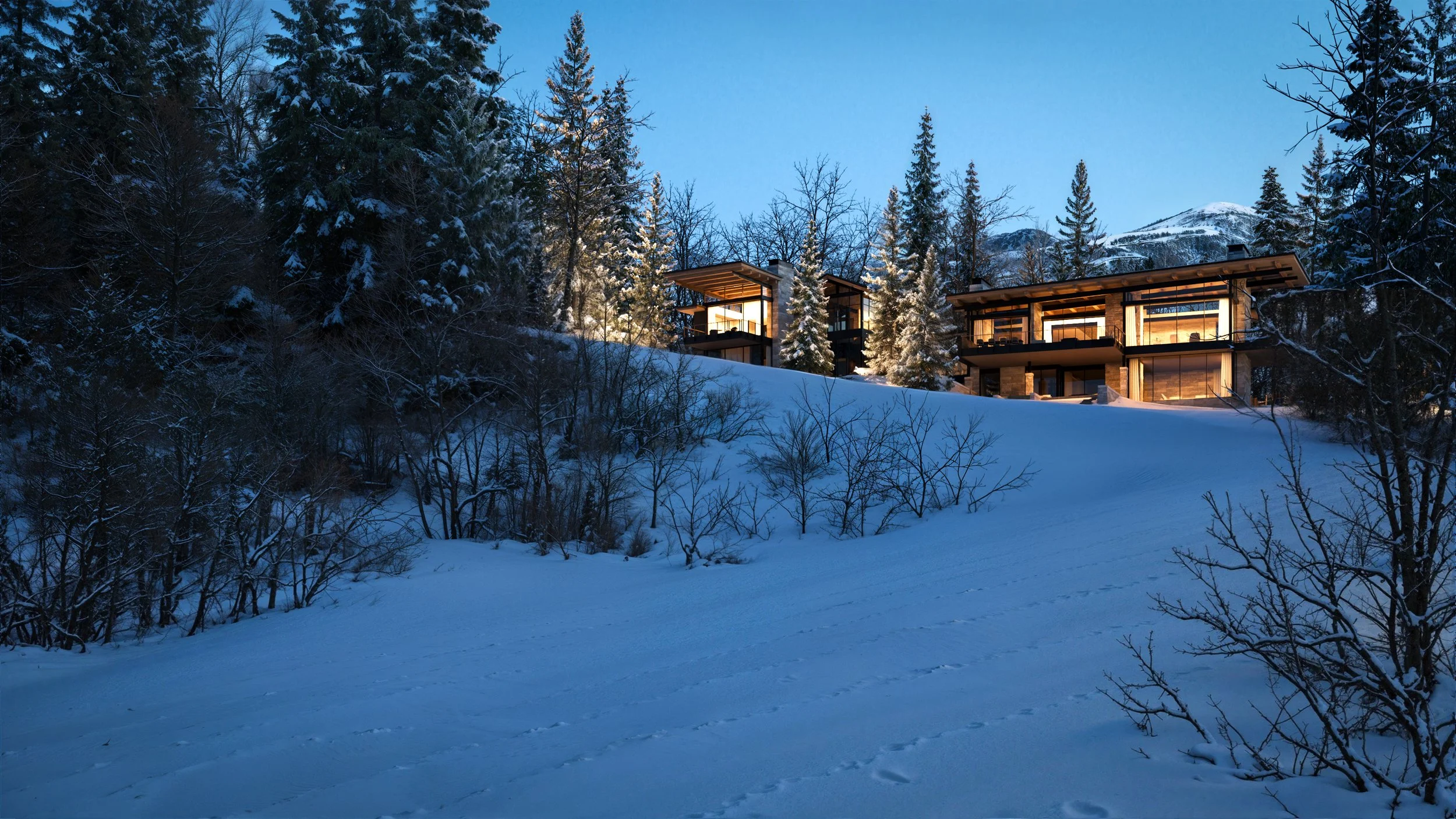 Modern house illuminated from inside, situated on a snow-covered hilltop surrounded by pine trees with snow, in a mountainous winter landscape at dusk.