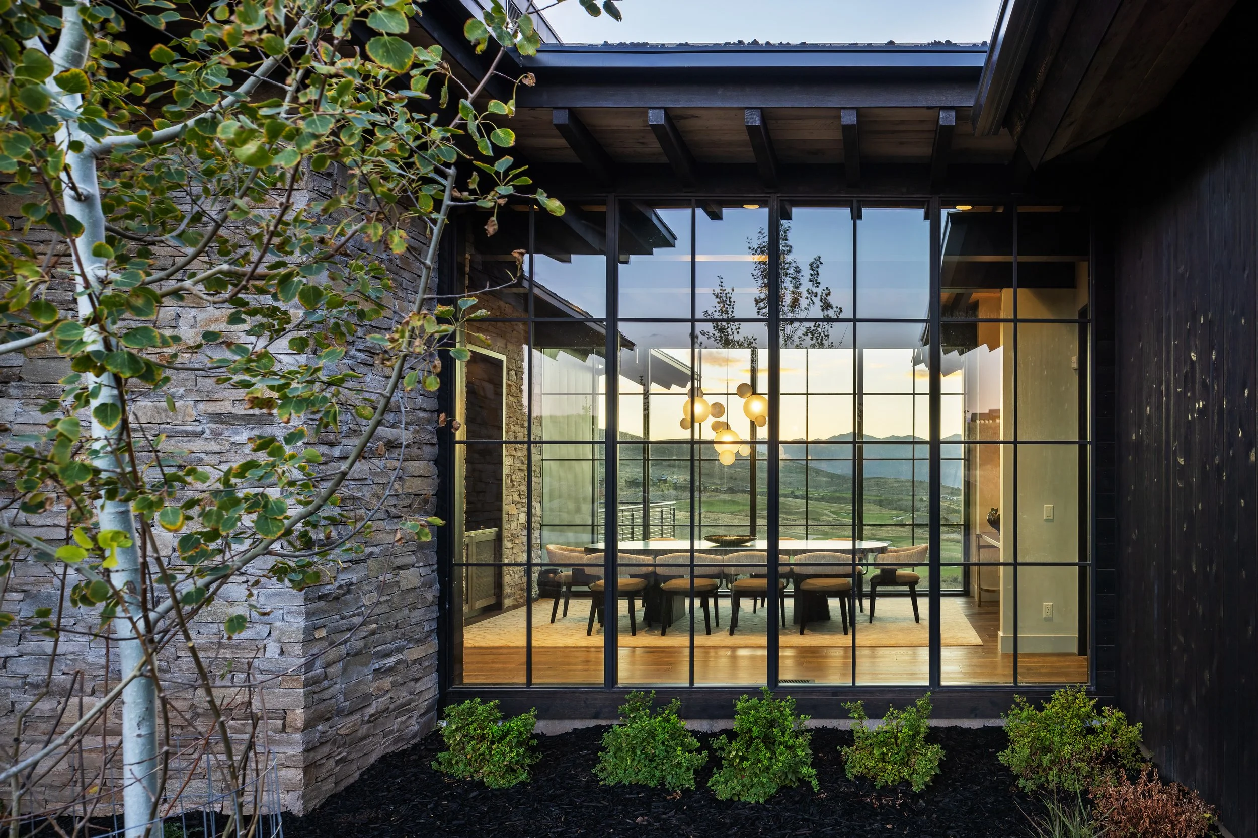 View through large glass windows of a dining room with a wooden table and chairs, modern chandelier, overlooking green hills at sunset, exterior includes stone and dark wood walls with landscaped garden.