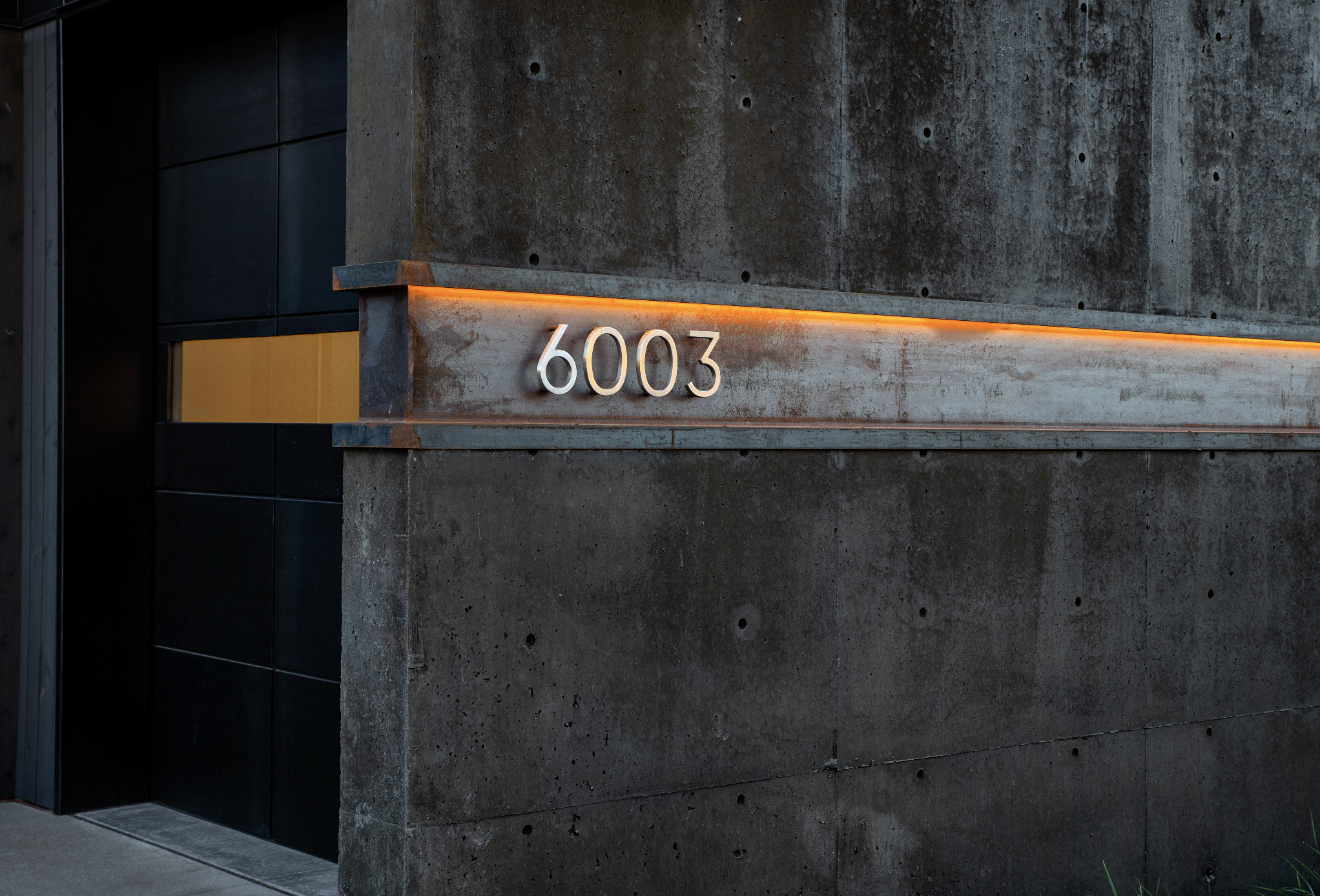 Building exterior with concrete wall and black tiles, modern house number sign "6003" on a metal strip with an orange light accent.