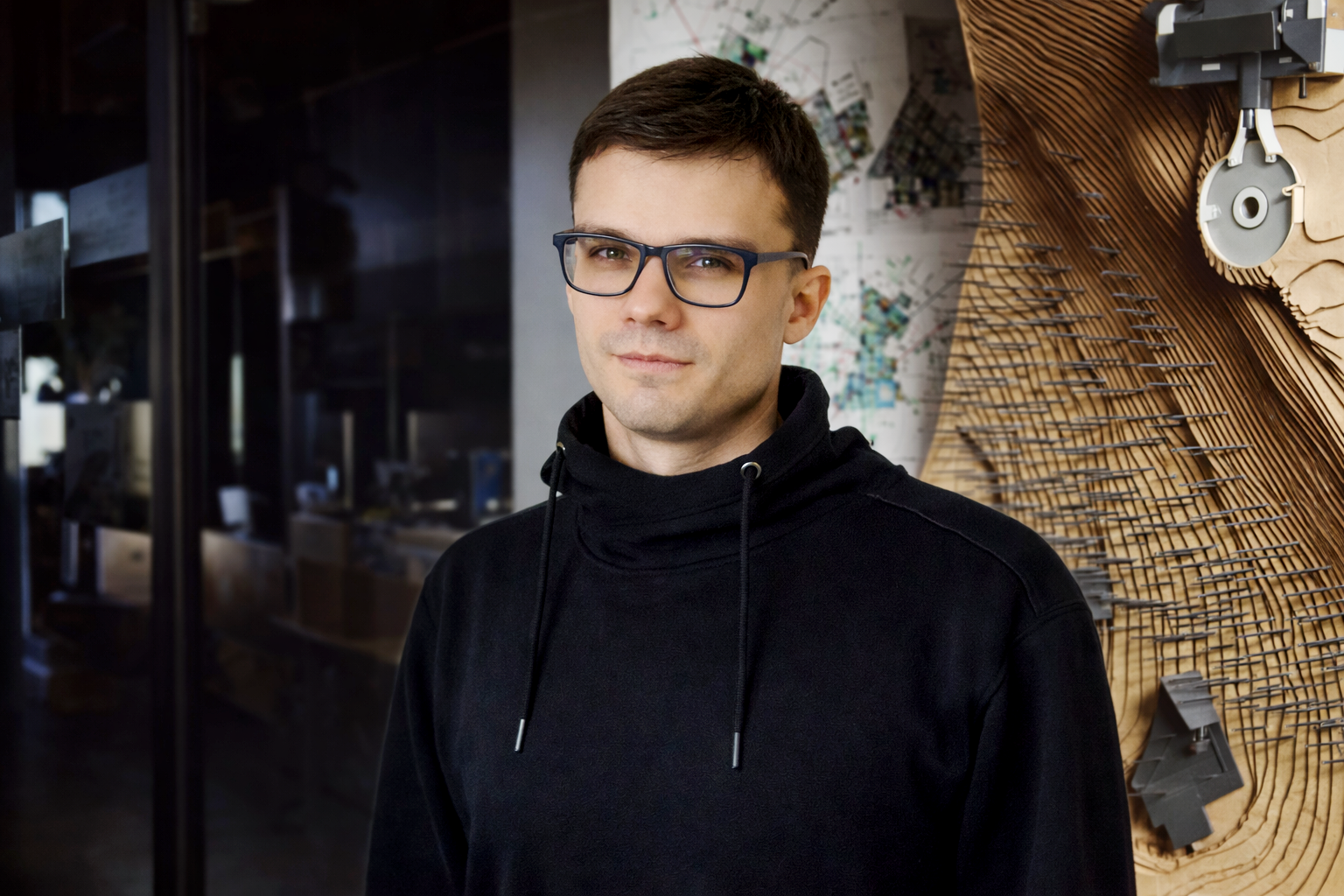 A young man with short brown hair and glasses, wearing a black hoodie, standing in a workshop or studio with wooden and mechanical tools in the background.