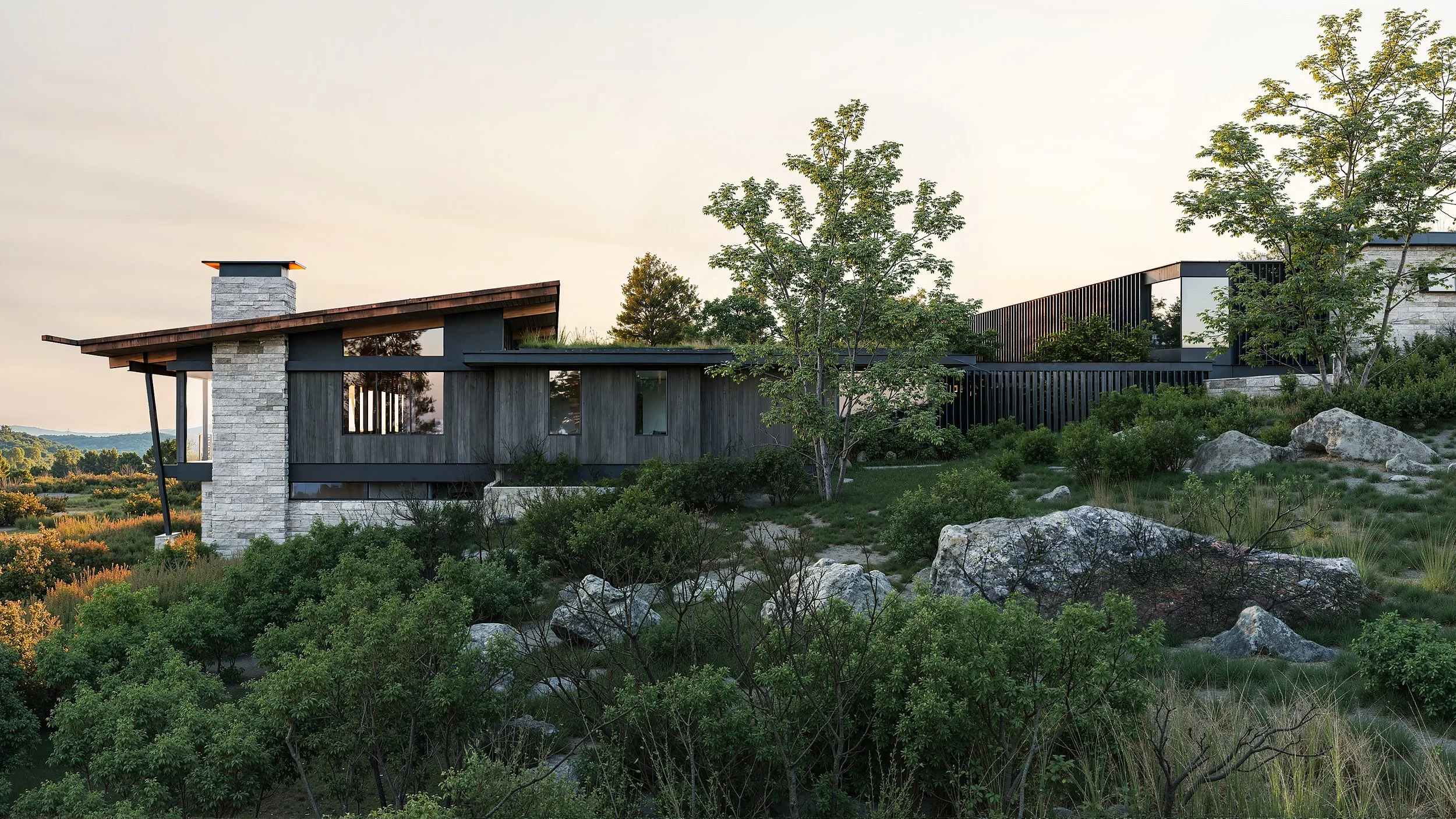 Modern house on a hillside with large rocks, trees, and greenery, under a clear sky during sunset.