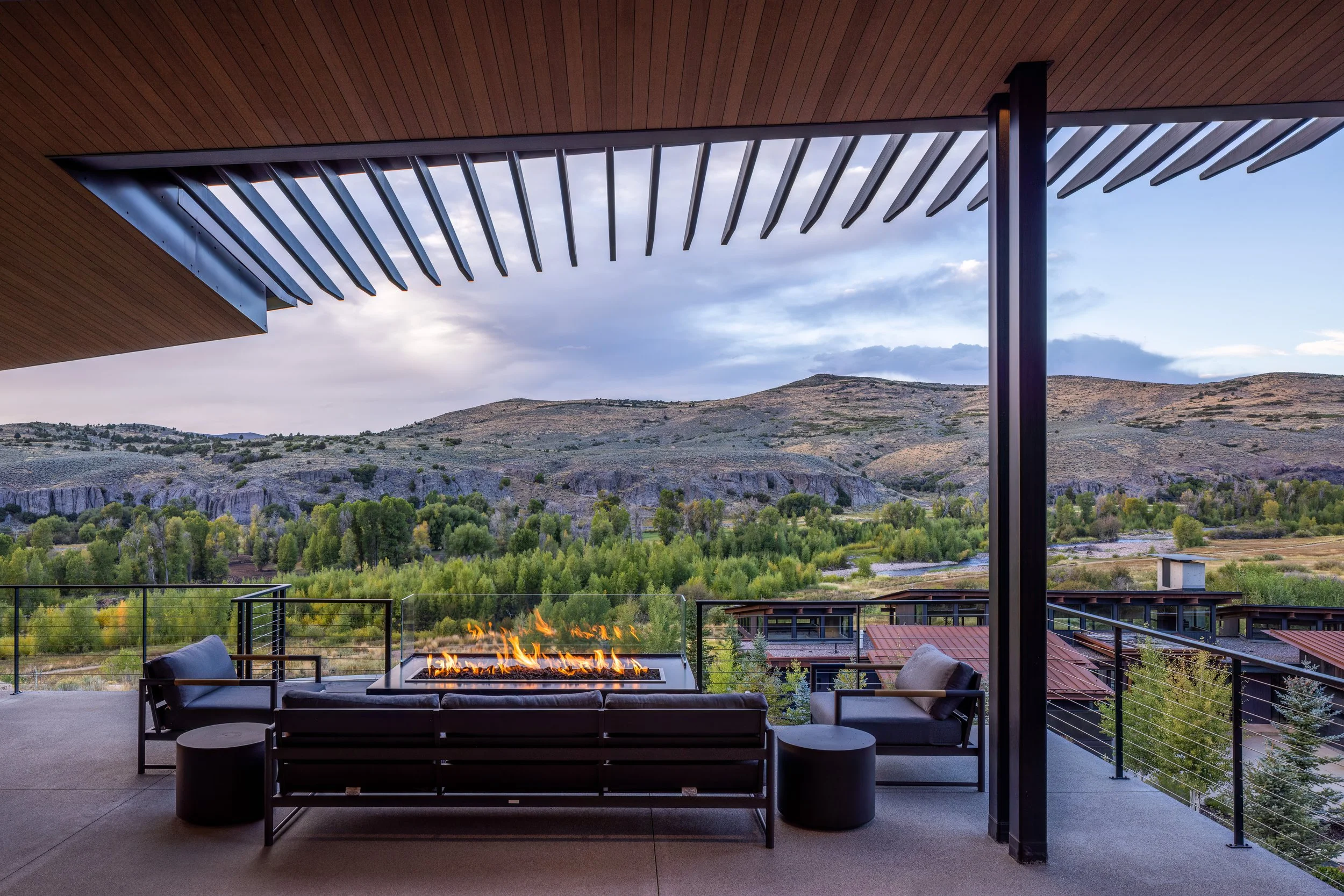 Modern balcony with outdoor seating and fire feature overlooking green hills and mountains under a cloudy sky.