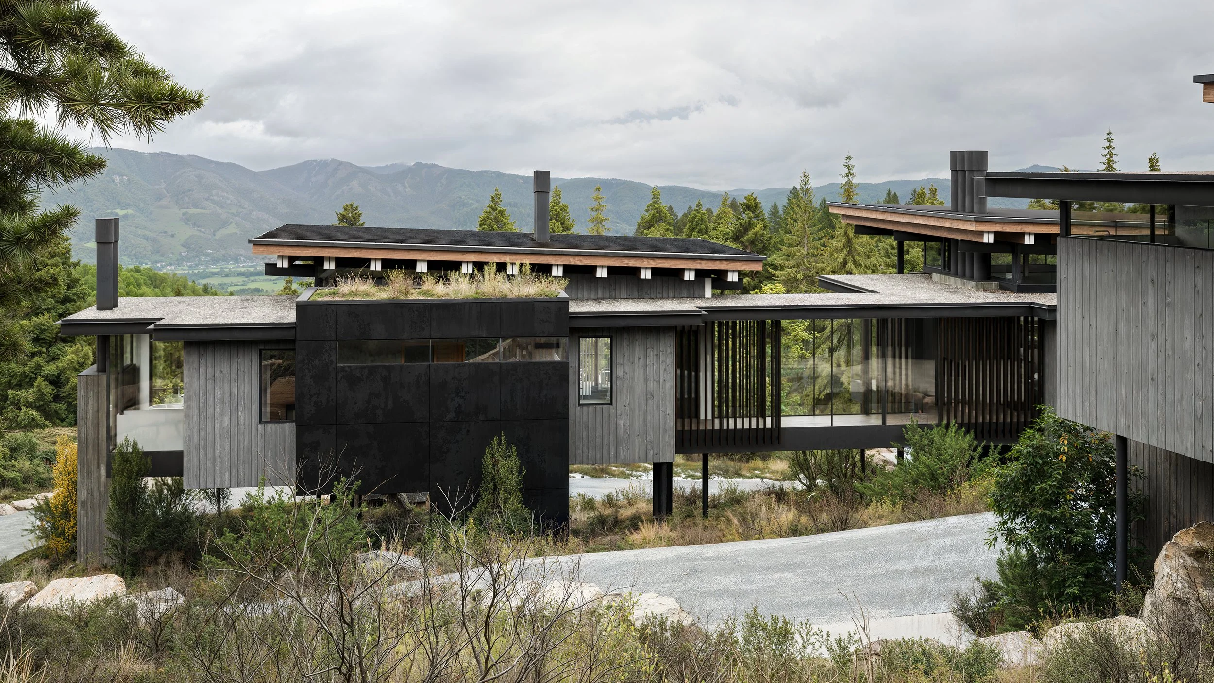 Modern house with black and gray exterior, large glass windows, surrounded by trees and mountains in the background.