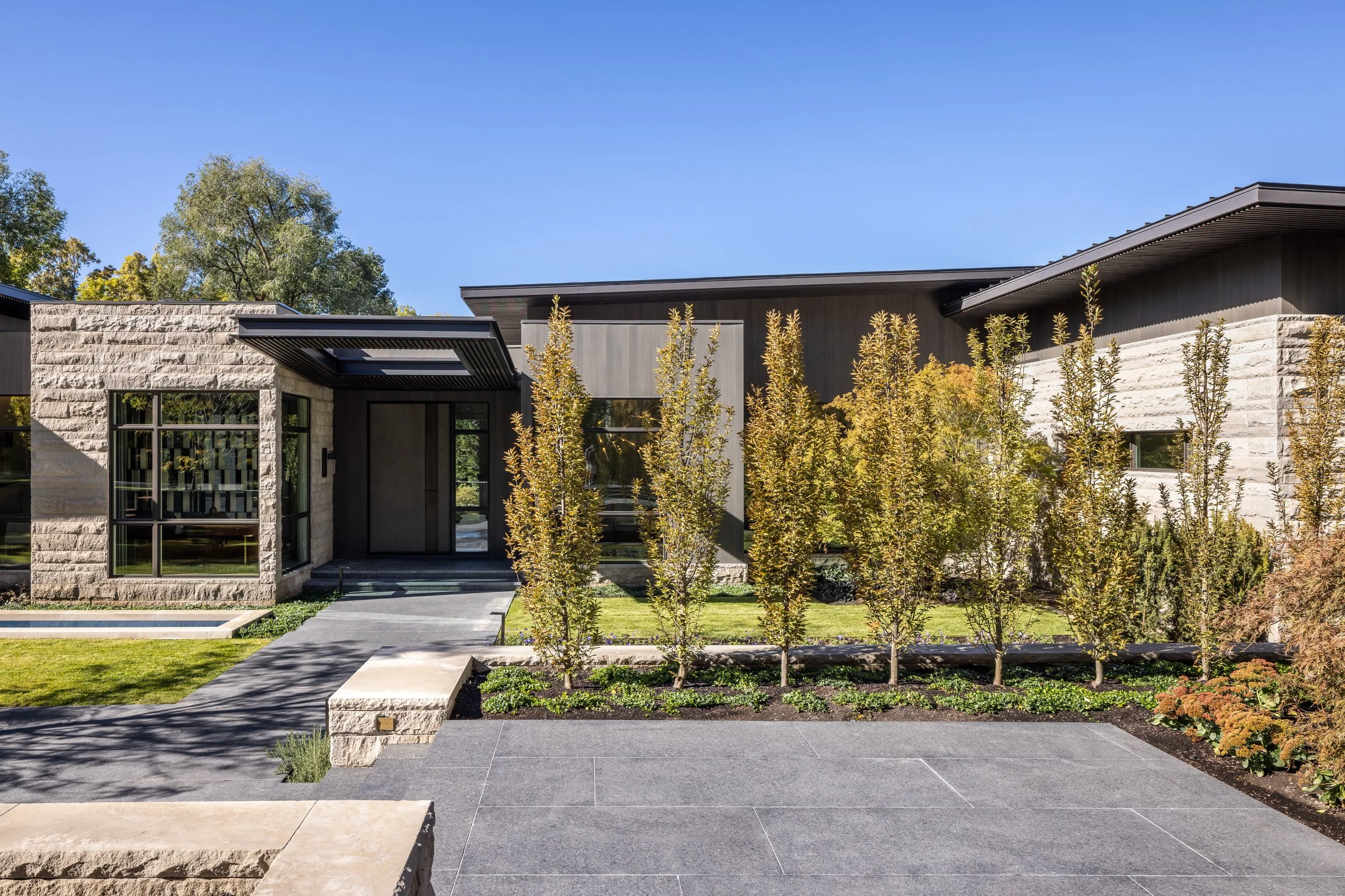 Modern house with stone and wood exterior, large windows, a small landscaped front yard with a row of tall plants, a concrete walkway, and a bright blue sky.