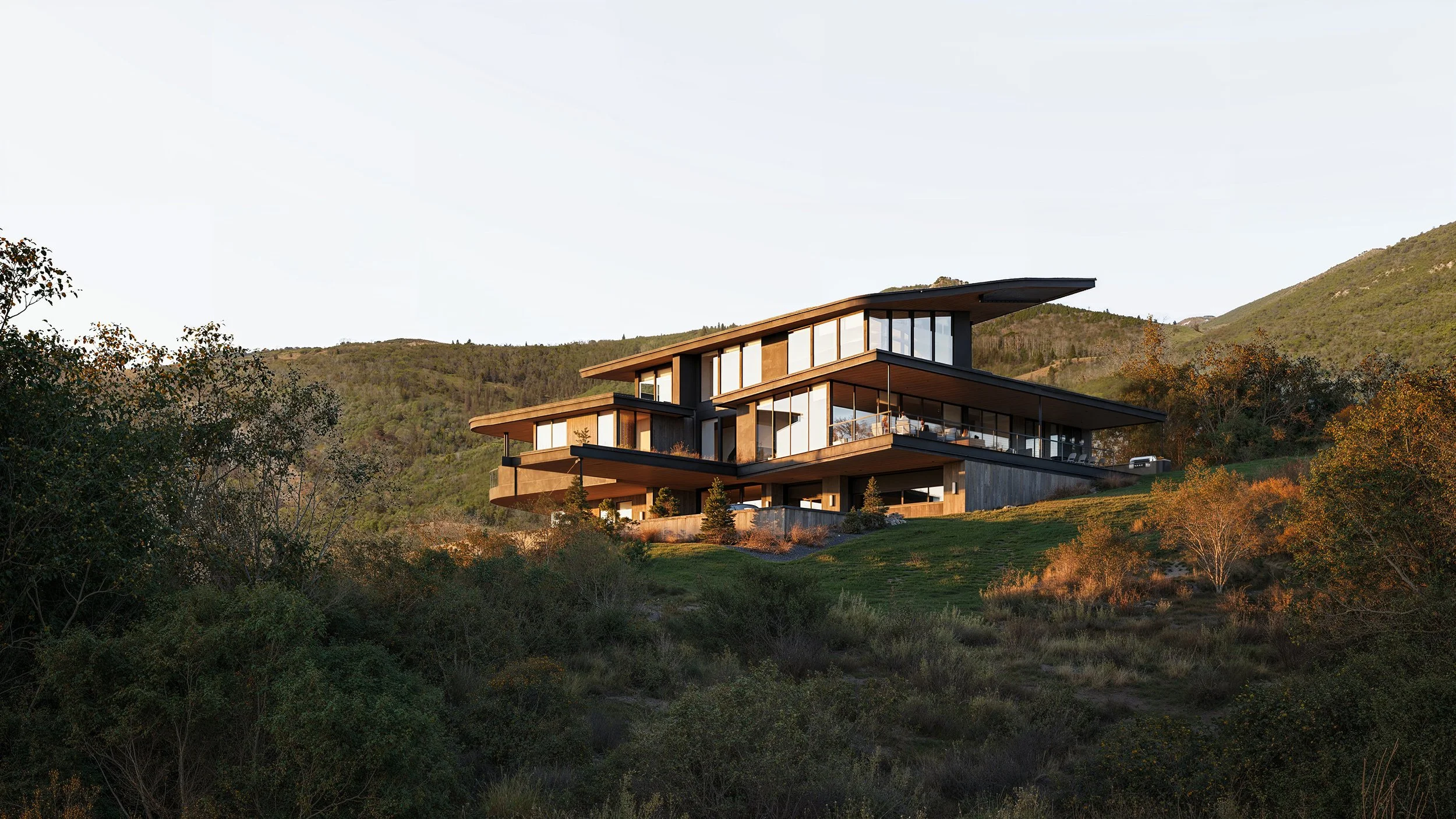 Modern house on a hillside with large glass windows, multiple levels, and surrounded by trees and mountains in the background during sunset.