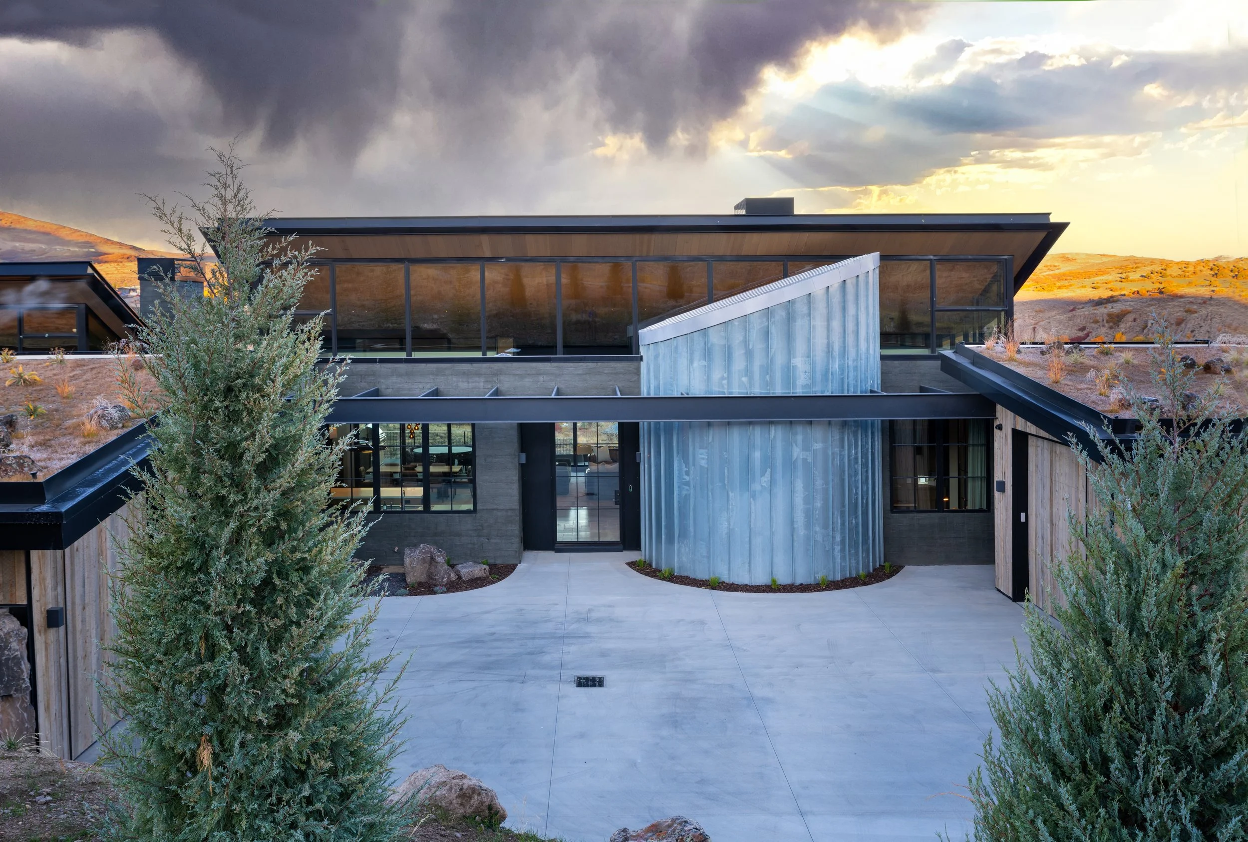 Modern house with a flat roof, large windows, including a rounded metal siding section, set in a natural landscape with hills and a stormy sky in the background.