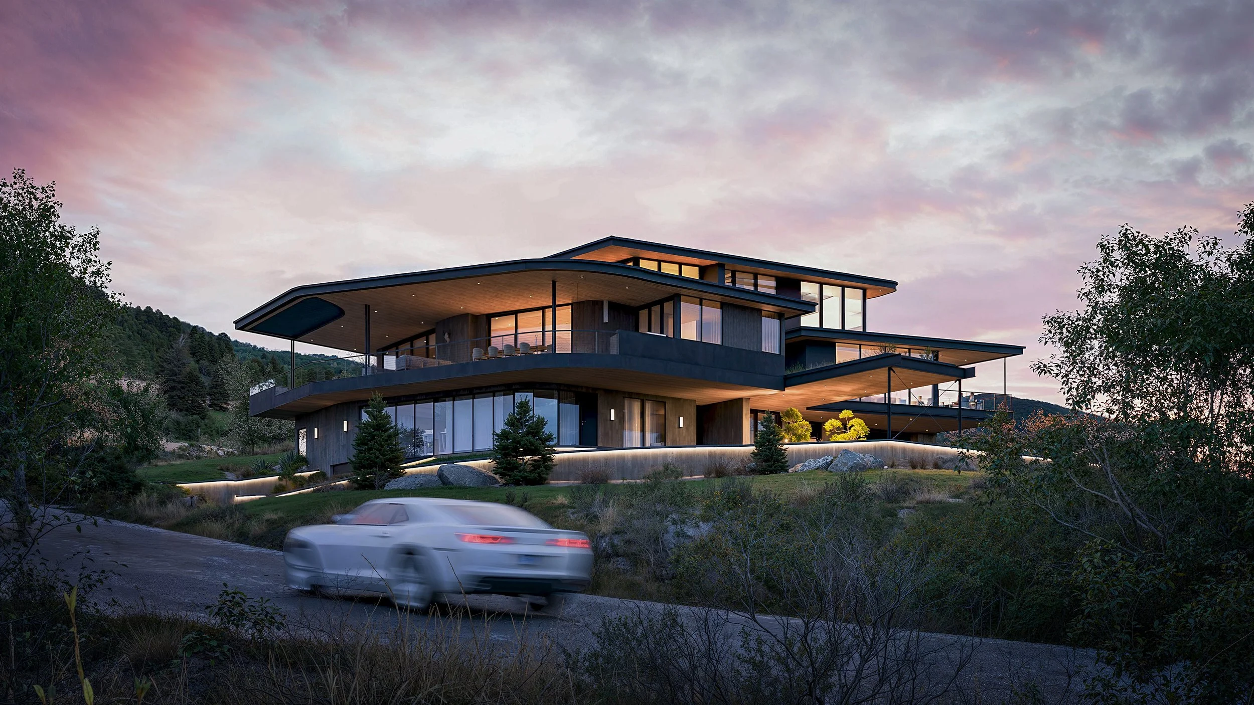 Modern multi-story house with large glass windows, situated on a hillside, with a car driving by on a winding road during sunset.