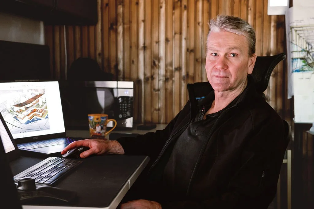 An older man with gray hair sitting at a desk in front of a computer with architectural plans on the screen, in a wooden-paneled room, with a coffee mug nearby.