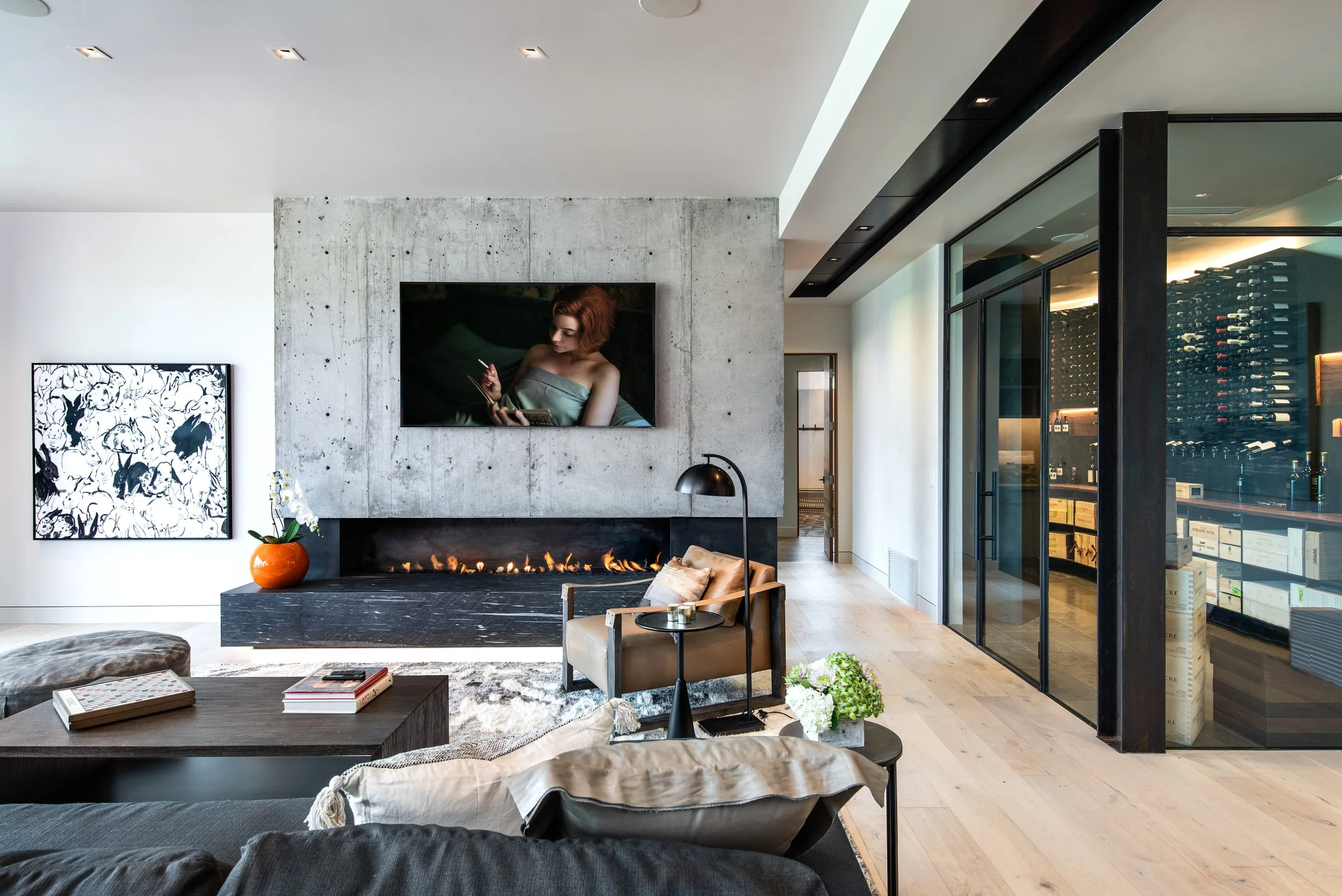 Modern living room with concrete wall, wall-mounted TV, fireplace, beige armchair, black side table with a lamp, artwork, and glass wine cellar.