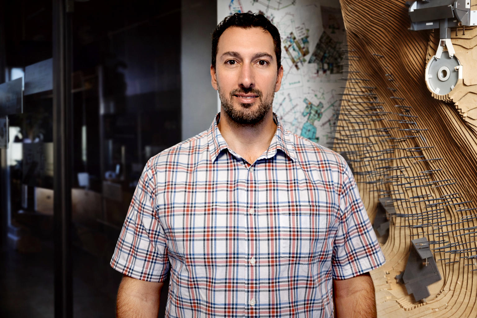 A man with dark hair and a beard wearing a short-sleeved, plaid button-up shirt standing in front of architectural models and drawings.