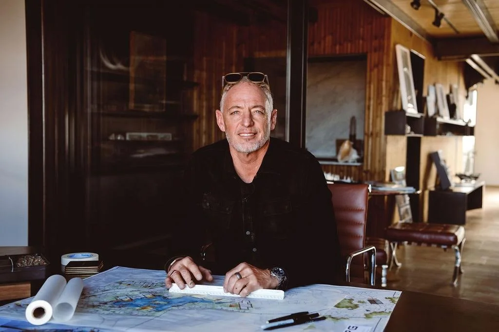 A middle-aged man with short gray hair and a beard, wearing sunglasses on his head and a black jacket, sitting at a table with maps and pens in a room with wooden walls and shelves, smiling at the camera.