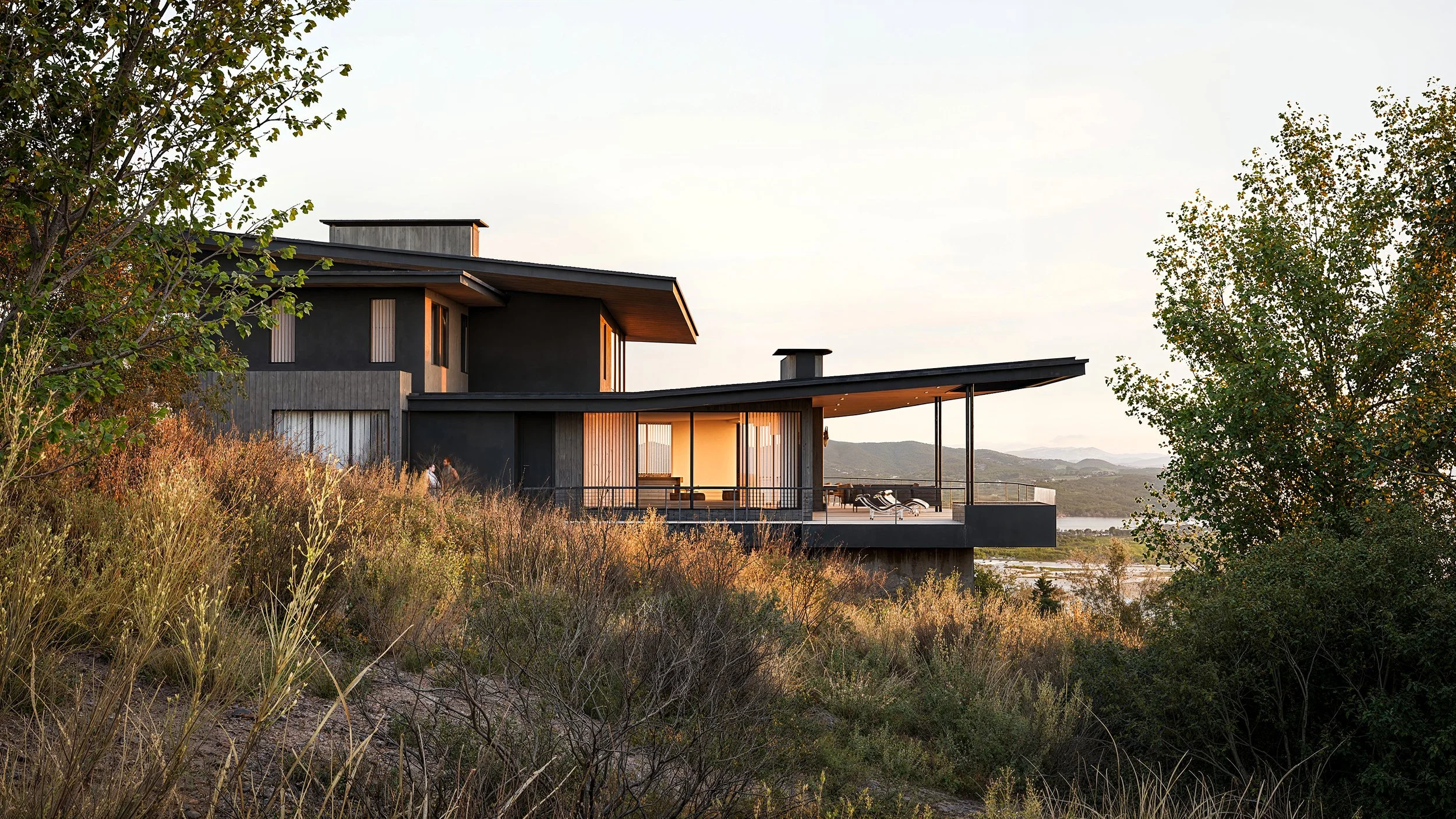 Modern house on a hill overlooking a body of water, with trees and dry grass in the foreground, and a mountain range in the distance during sunset.