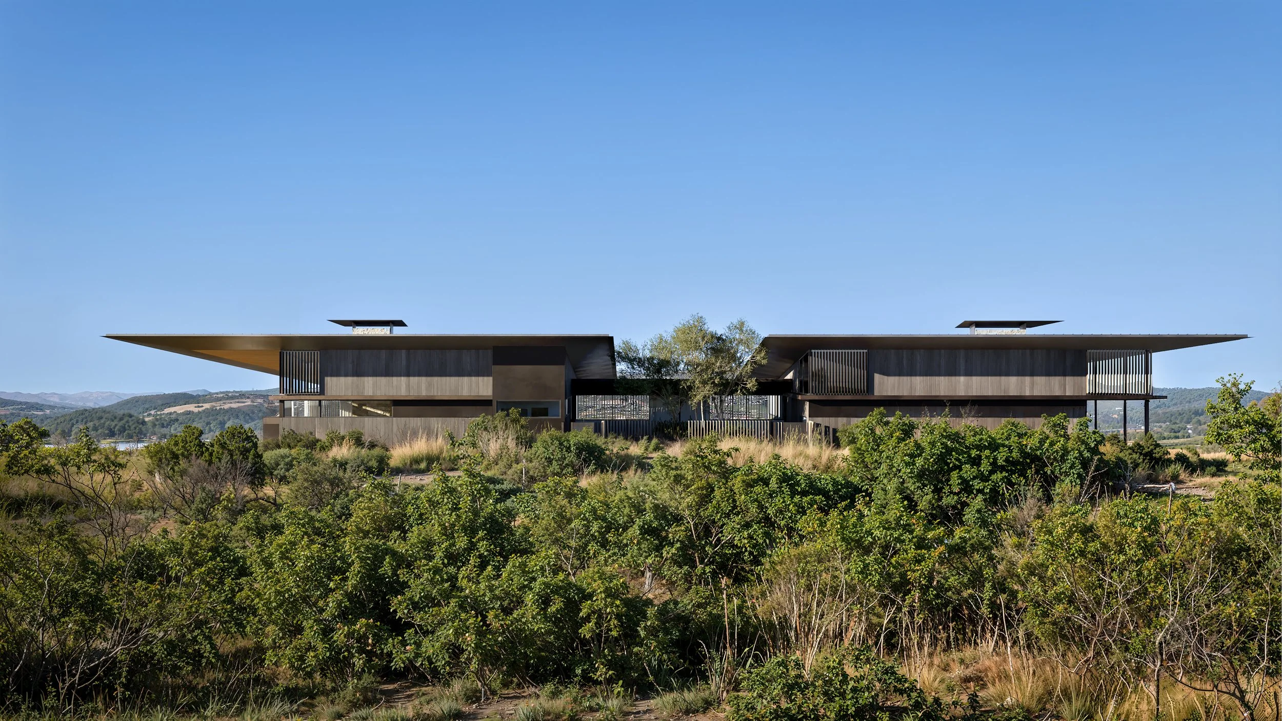 Modern two-story house with large, flat overhanging roof, surrounded by lush green vegetation, under a clear blue sky.