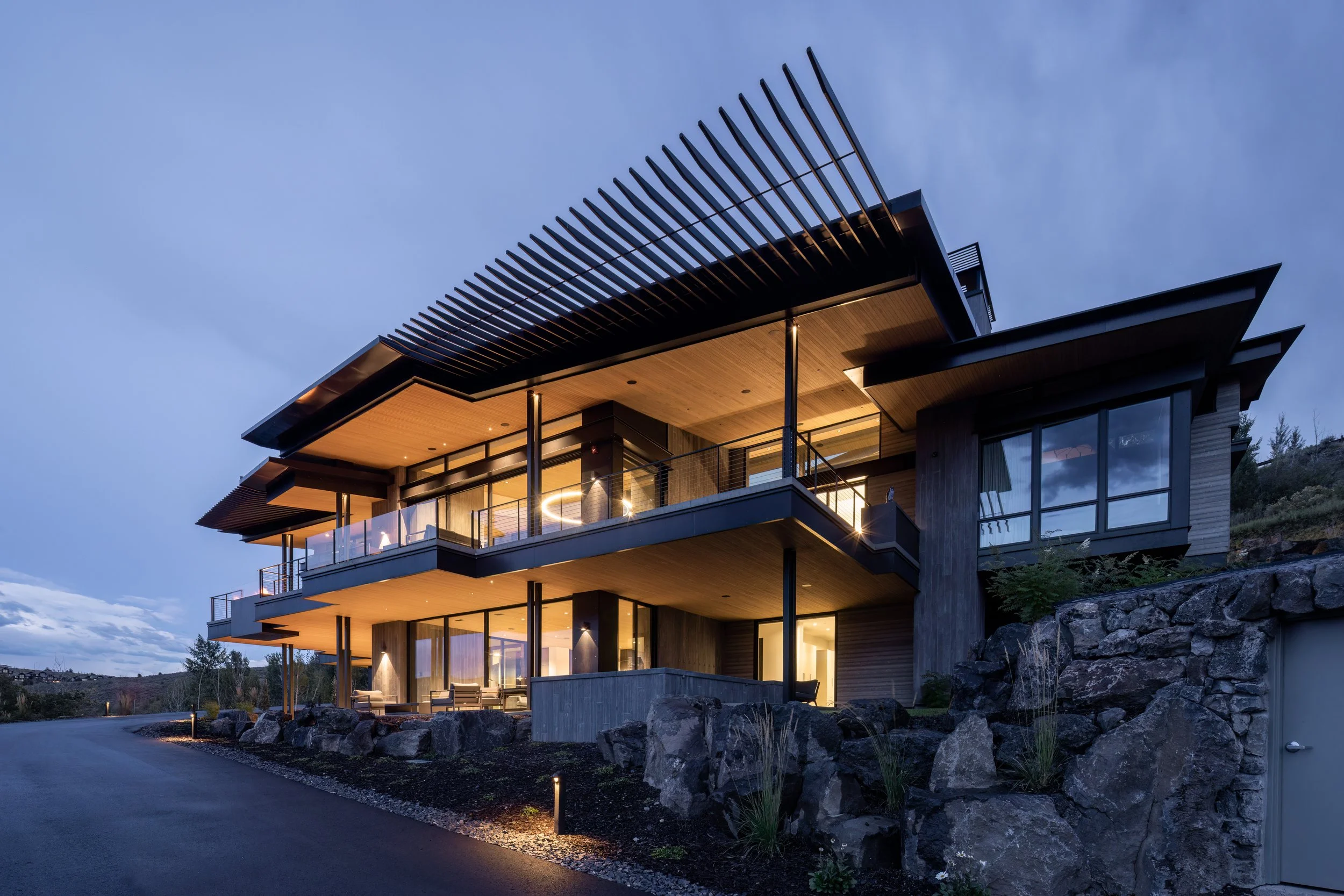 Modern multi-story house with glass walls and balconies, illuminated at dusk, built on a rocky landscape.
