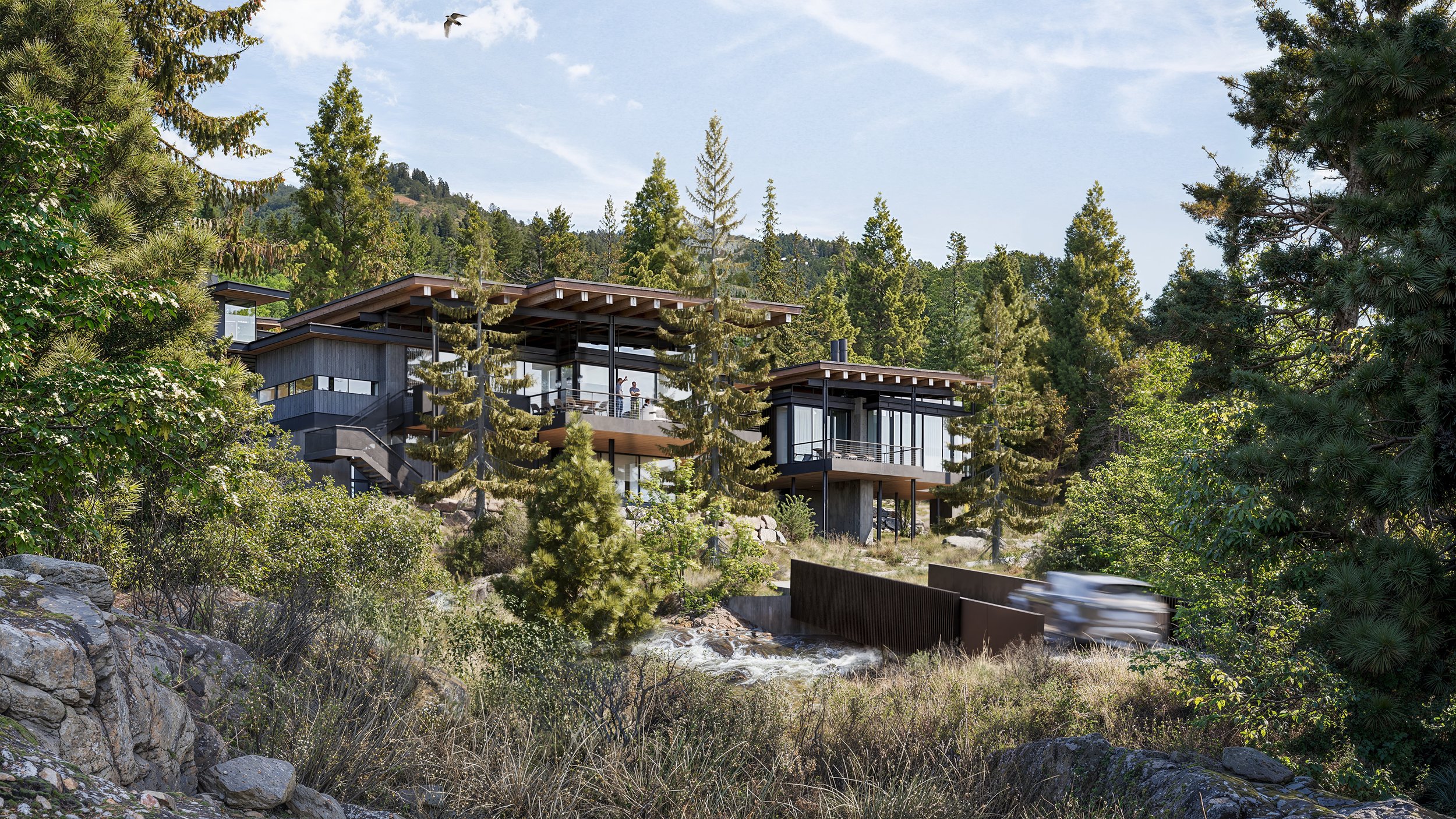 Modern house with large glass windows and multiple levels, situated on a hillside among tall pine trees, with a flowing stream and rocks in the foreground.
