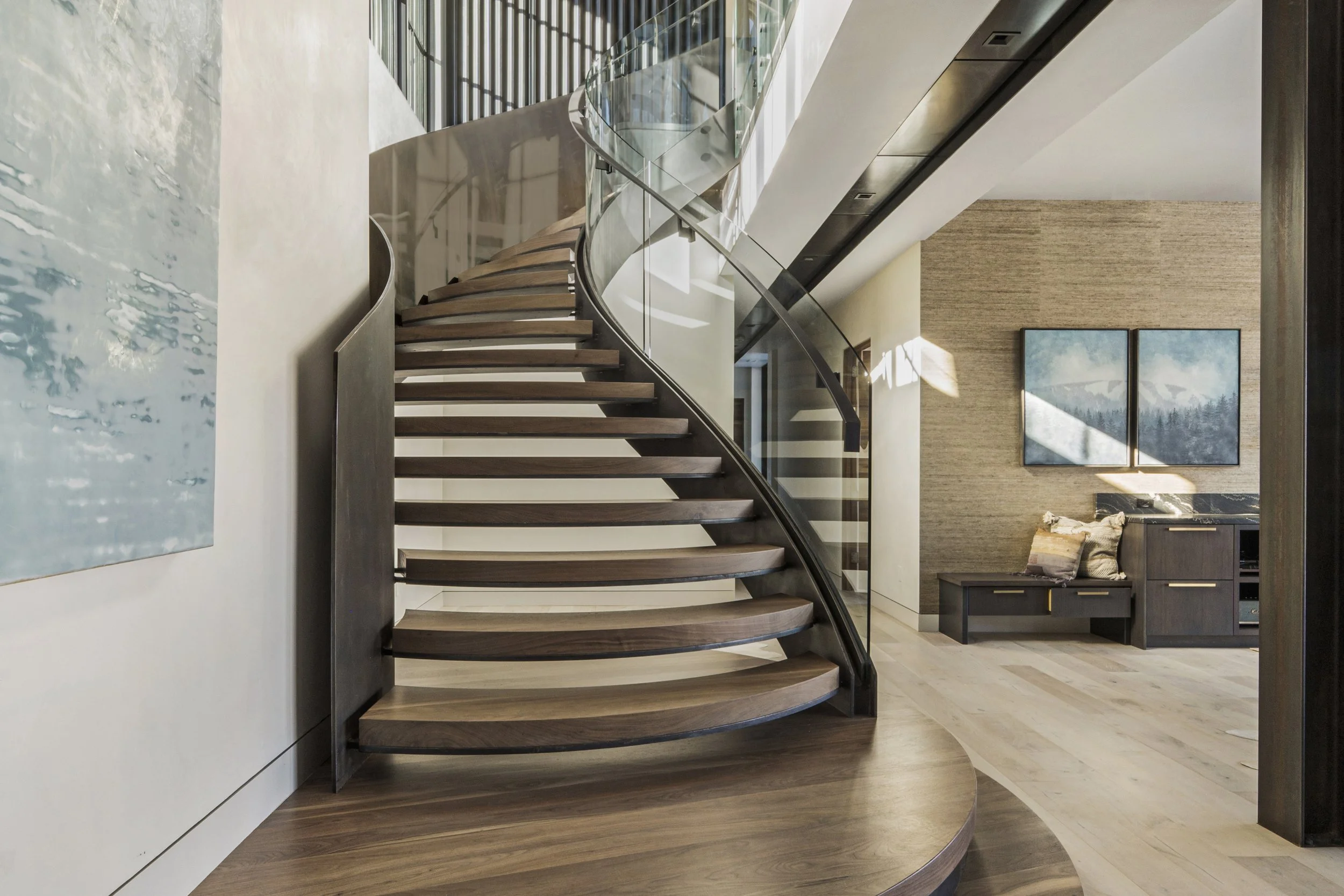 Modern interior space featuring a curved wooden staircase with glass railing, light wood flooring, a bench, wall art, and a textured wall in the background.