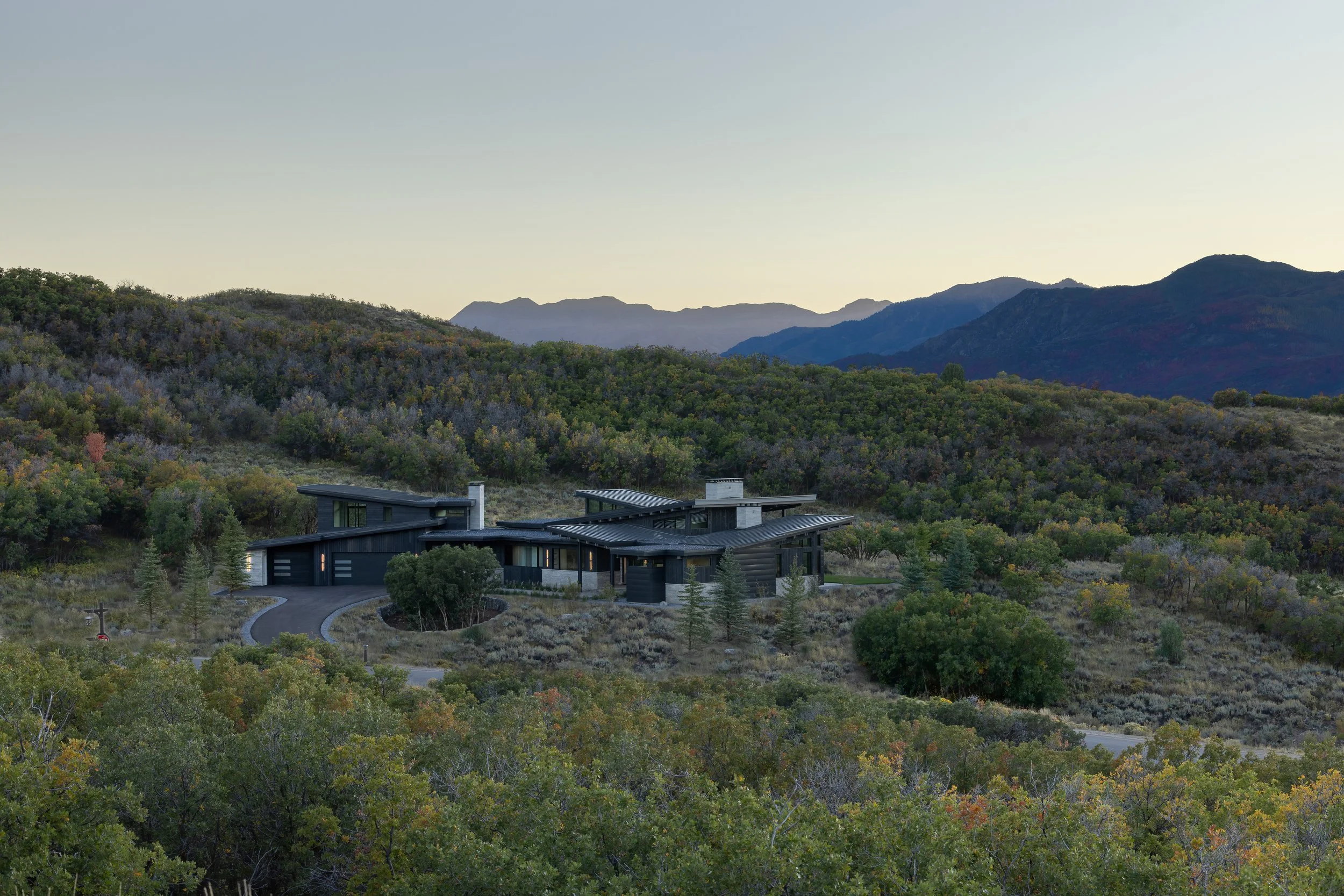 Modern black house with flat and slanted roofs, situated in a hilly landscape with various green and brown vegetation, mountains in the background, under a clear sky.