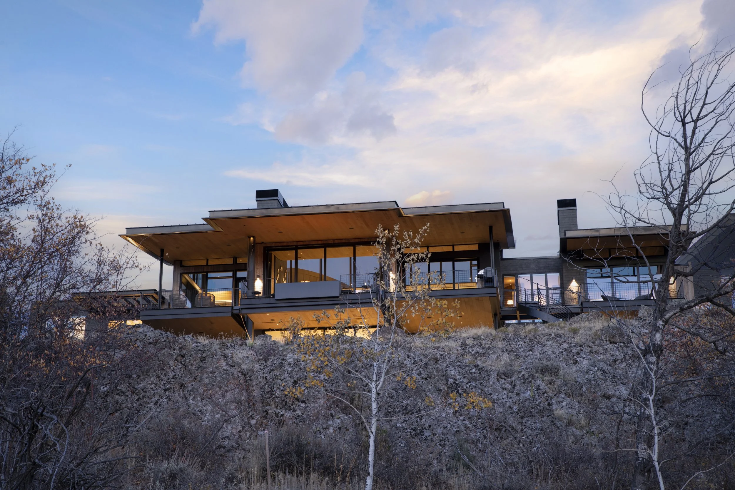 Modern house on a hilltop during sunset, with large windows and a wooden roof, surrounded by leafless trees and rocky landscape.