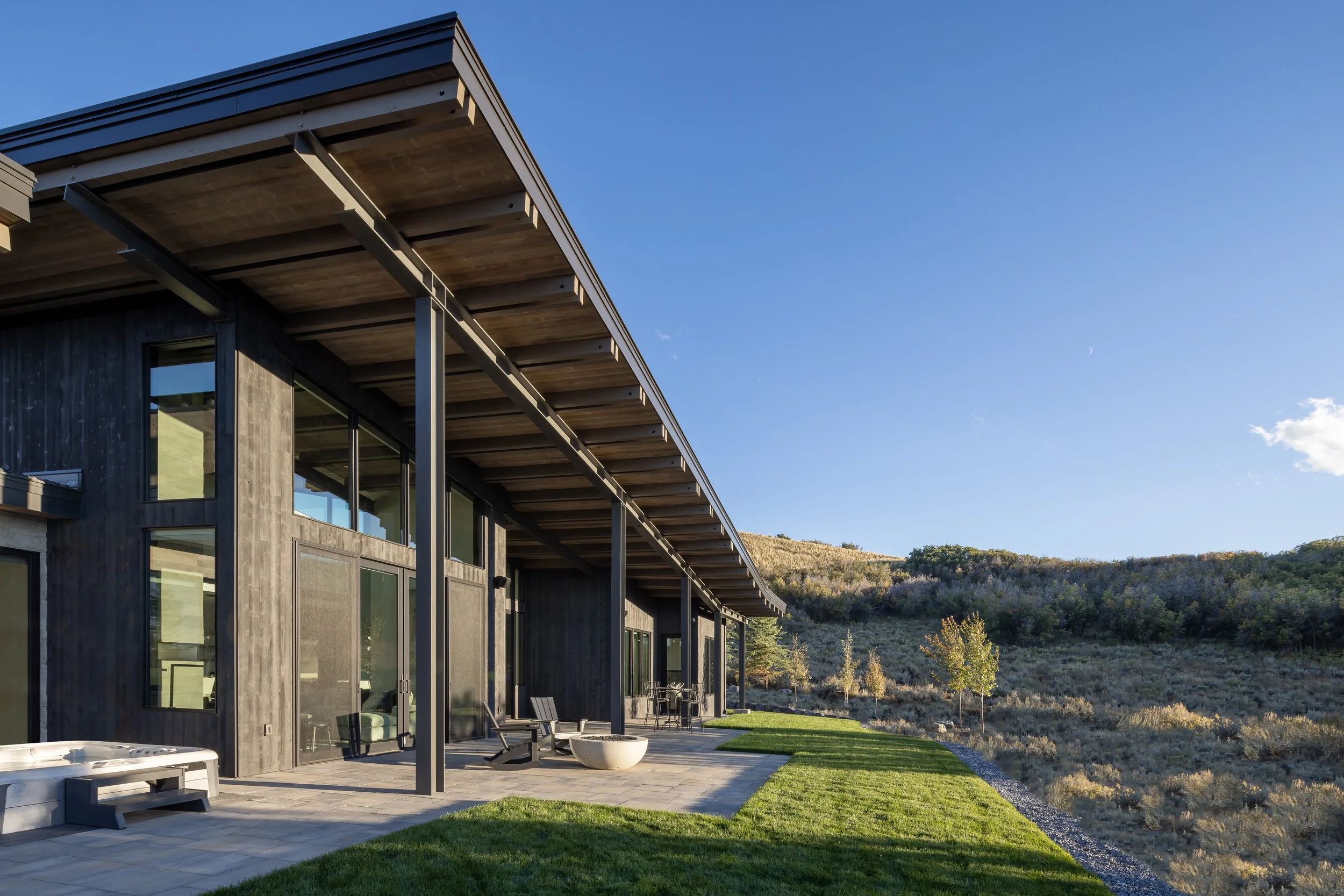 Modern house with a large patio and outdoor seating, overlooking a hill with sparse vegetation under a clear blue sky.