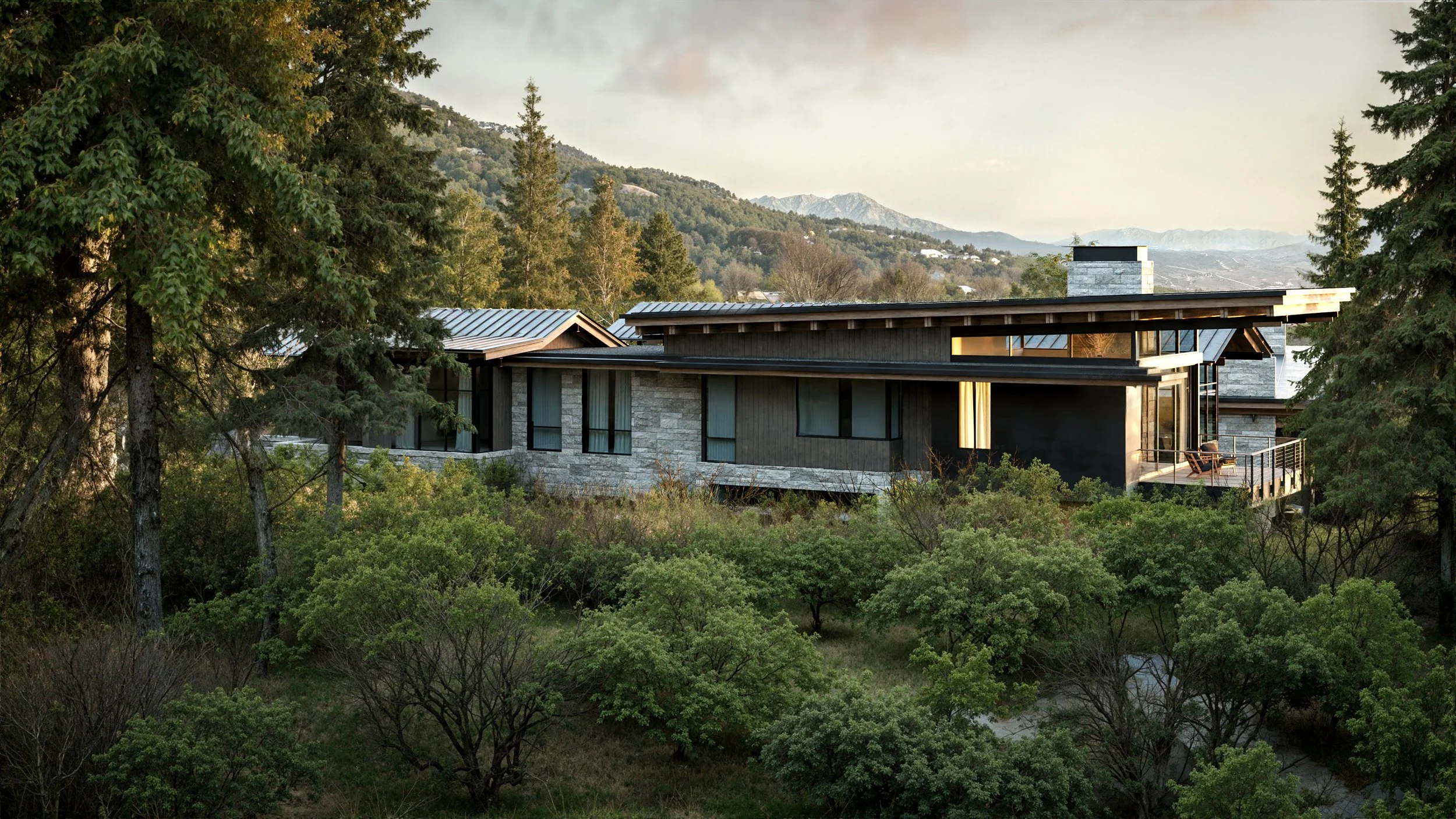 Modern house with flat roof, large windows, and stone exterior, situated among green trees and mountains in the background, with a balcony on the right side.