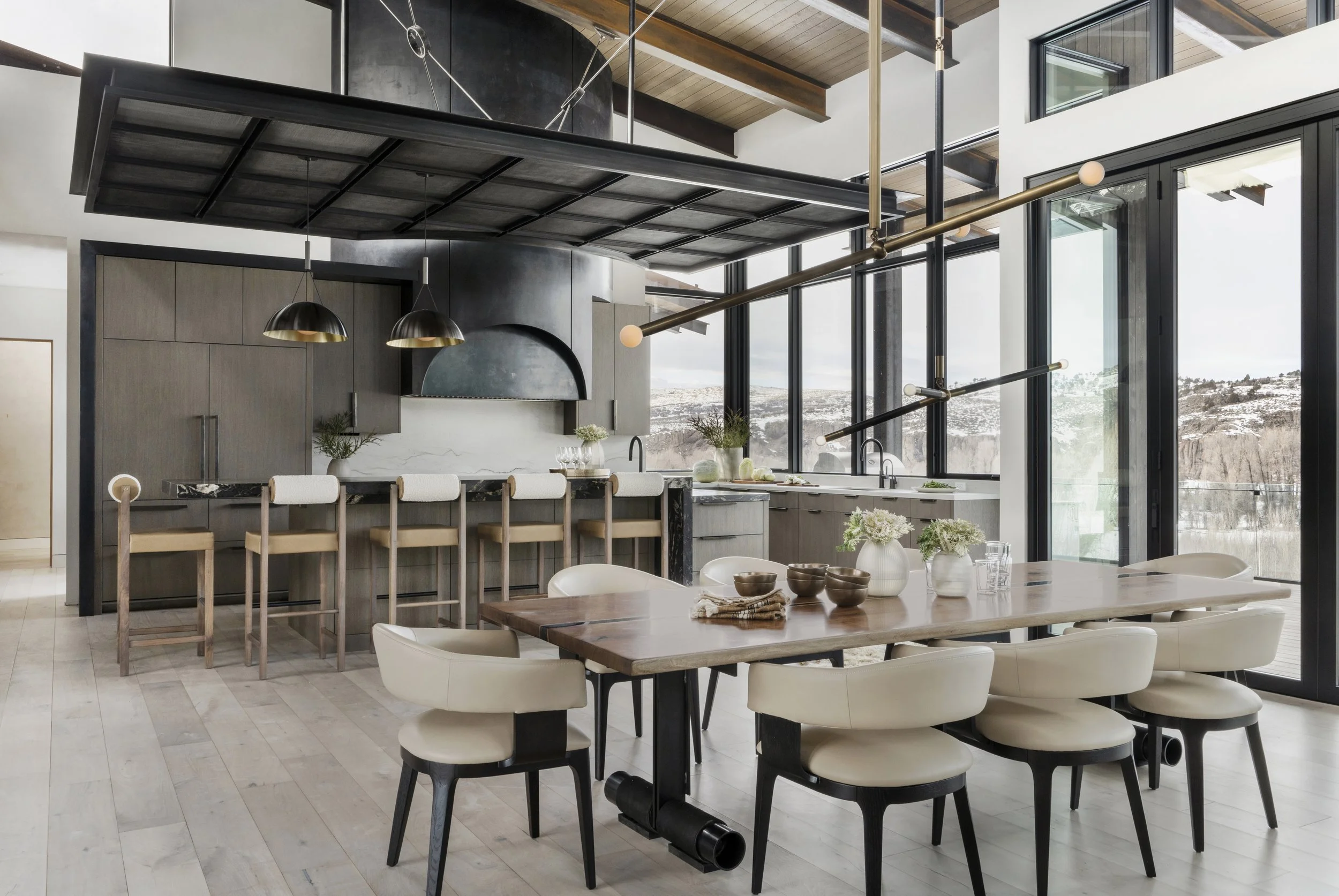 Modern open-concept kitchen and dining area with a large wooden table, white chairs, and a kitchen island with bar stools. Floor-to-ceiling windows offer a view of snowy landscape outside.