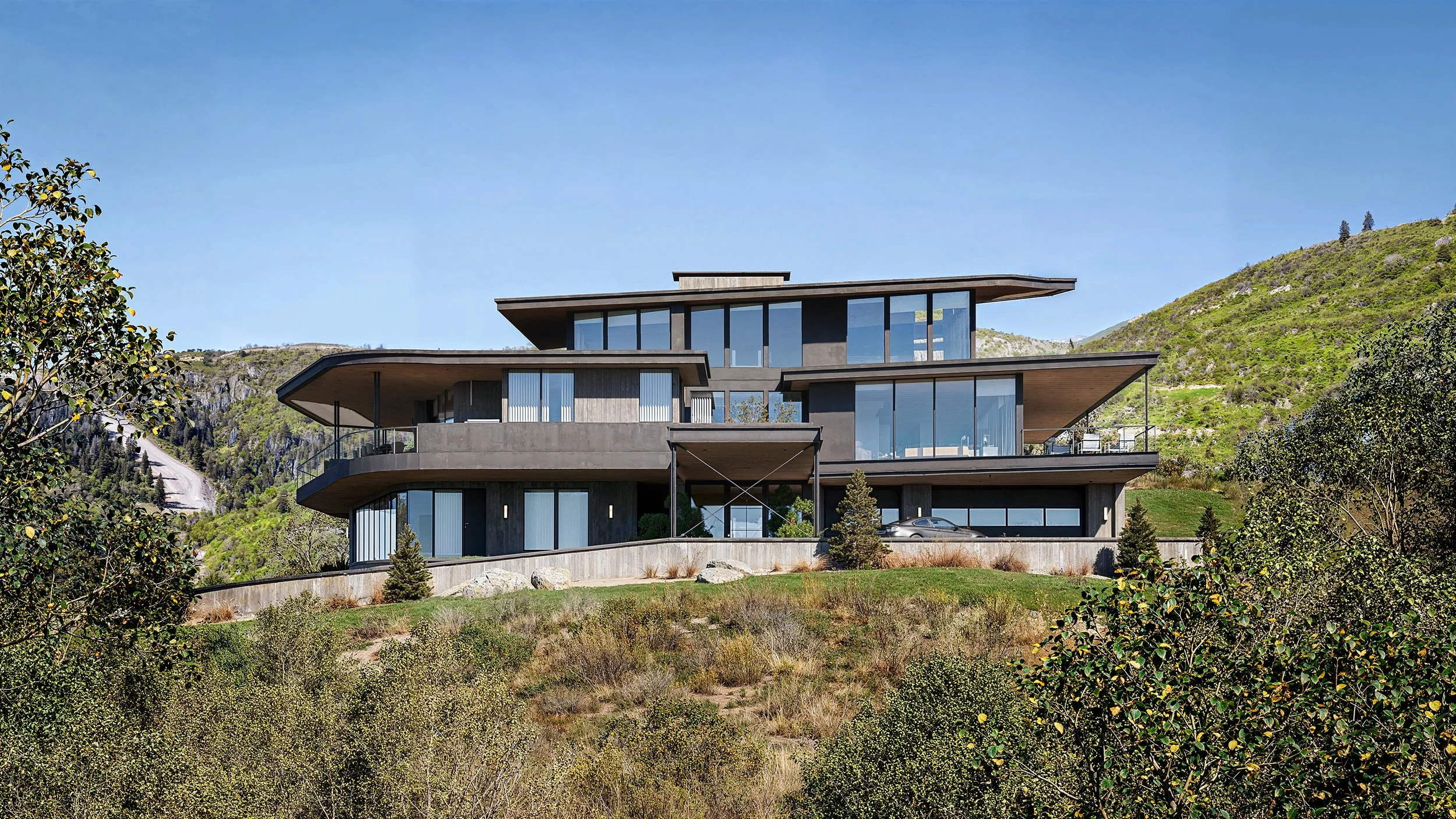 Modern multi-story house with large glass windows and flat, overlapping rooflines situated on a hillside surrounded by greenery and mountains.