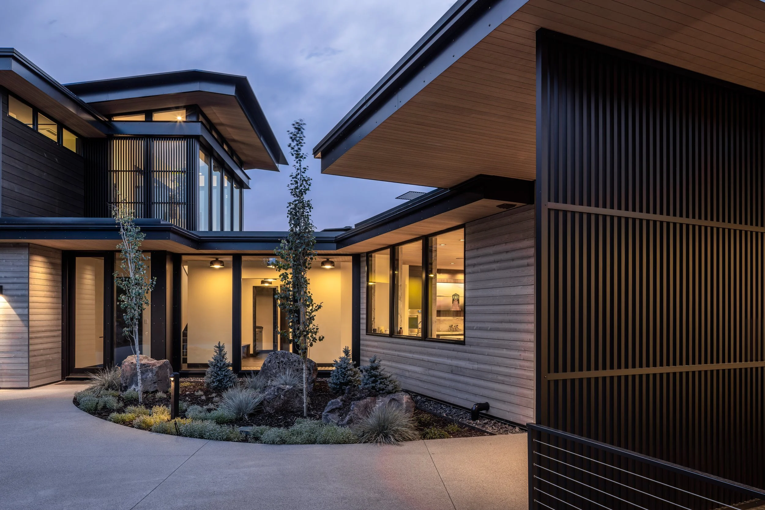Modern house exterior with large windows, wooden siding, and landscaped front yard during evening. Internal lights are on, illuminating the entryway and interior spaces.