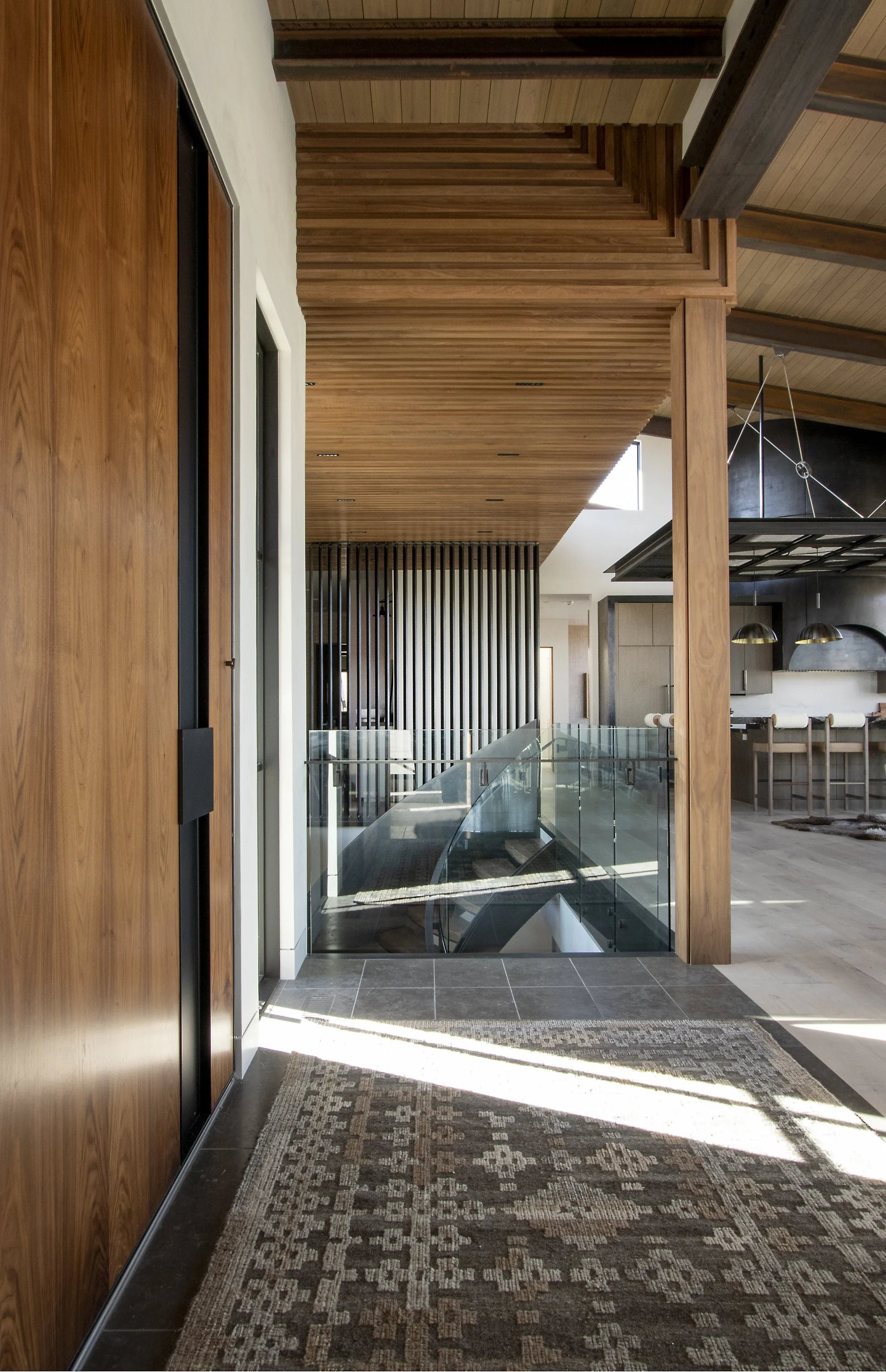 Interior view of a modern home with wooden ceiling and walls, glass railing overlooking a staircase, and a kitchen area with bar stools in the background.