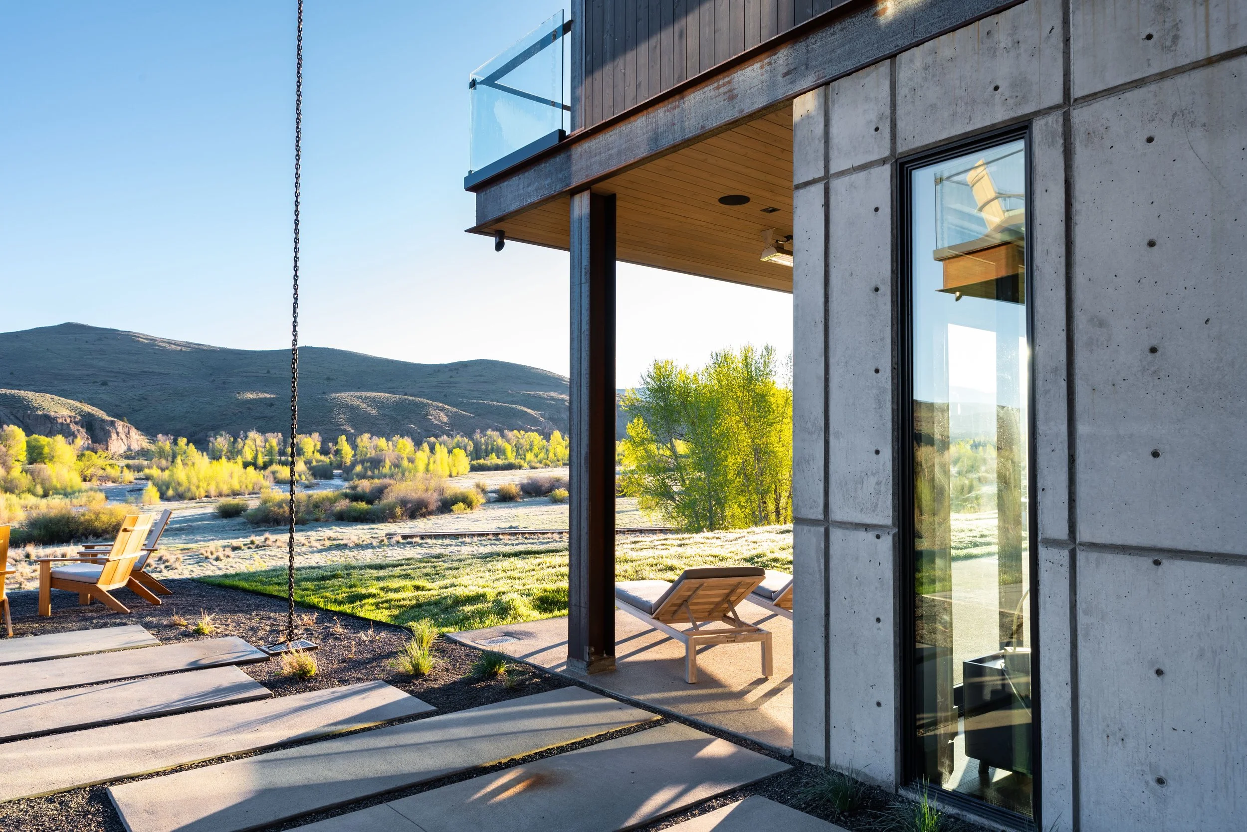 Modern house exterior with concrete wall, glass window, wooden ceiling, and outdoor seating area, overlooking a scenic landscape with hills, trees, and a river during daytime.