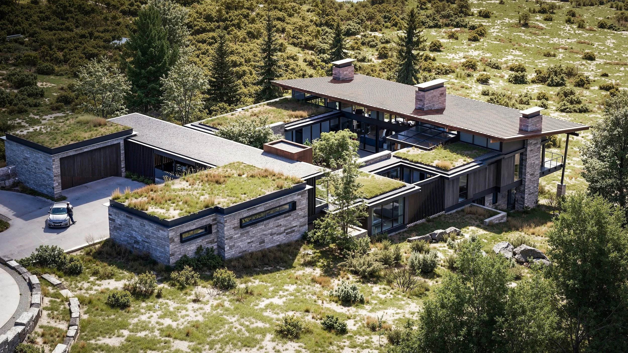 A modern house with flat, green roofs, surrounded by trees and natural landscape, with a driveway and parked car.