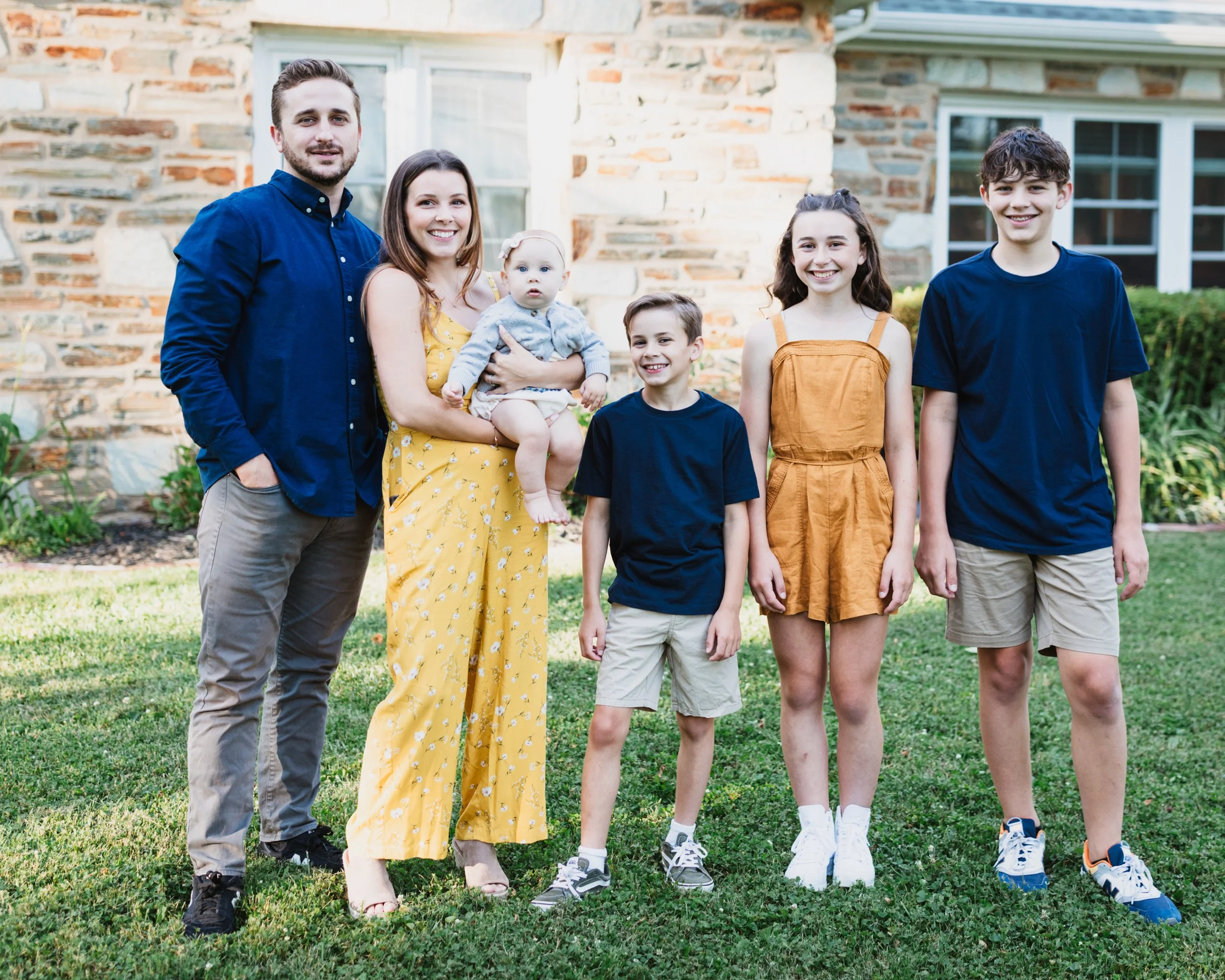 A family of six standing on a lawn in front of a brick house. The family includes a man, a woman holding a baby girl, and three children. Everyone is smiling and dressed casually.