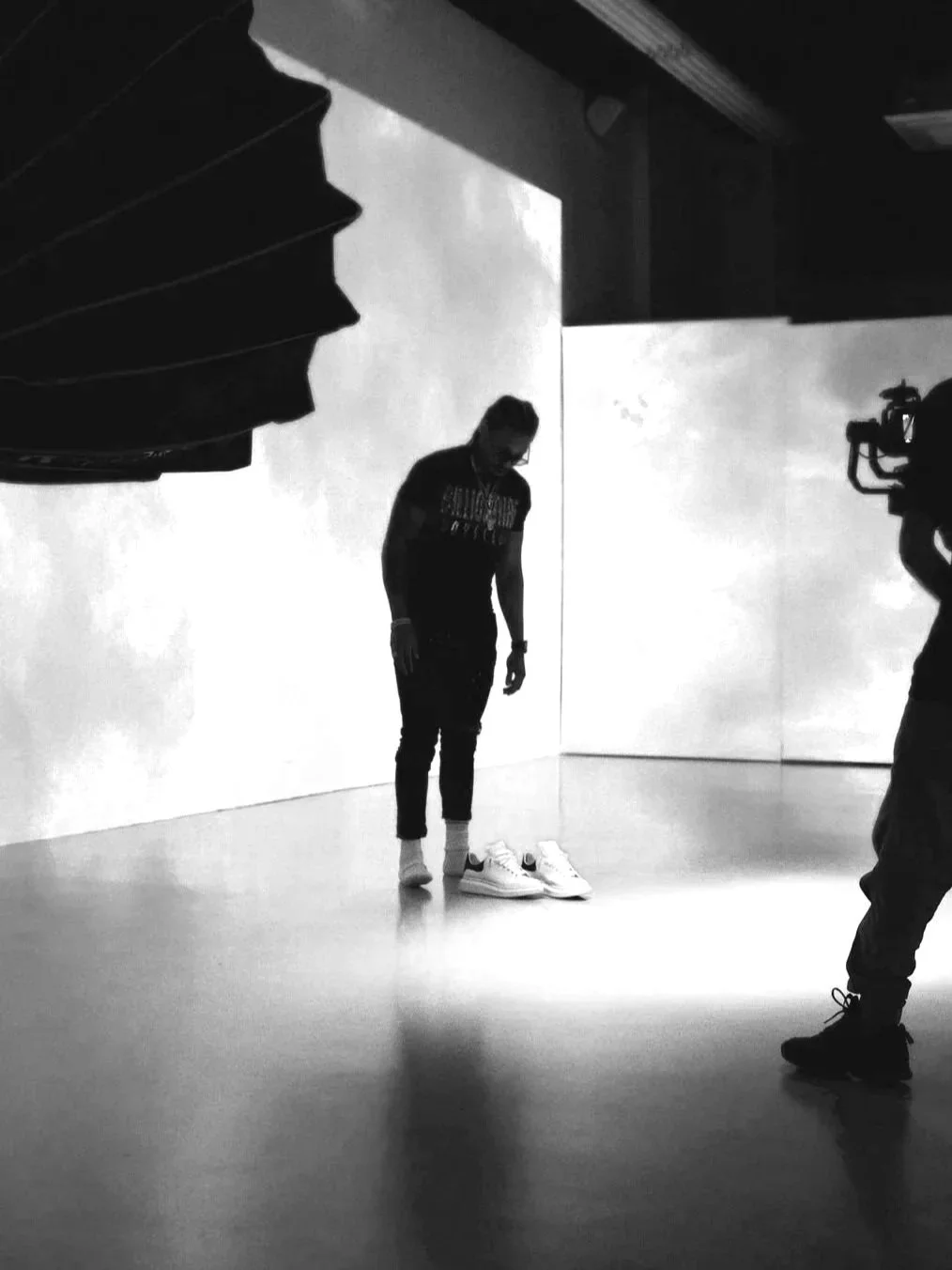 Person standing on a plain floor, looking down at a pair of white sneakers, with a photographer filming in front and a large backdrop behind.