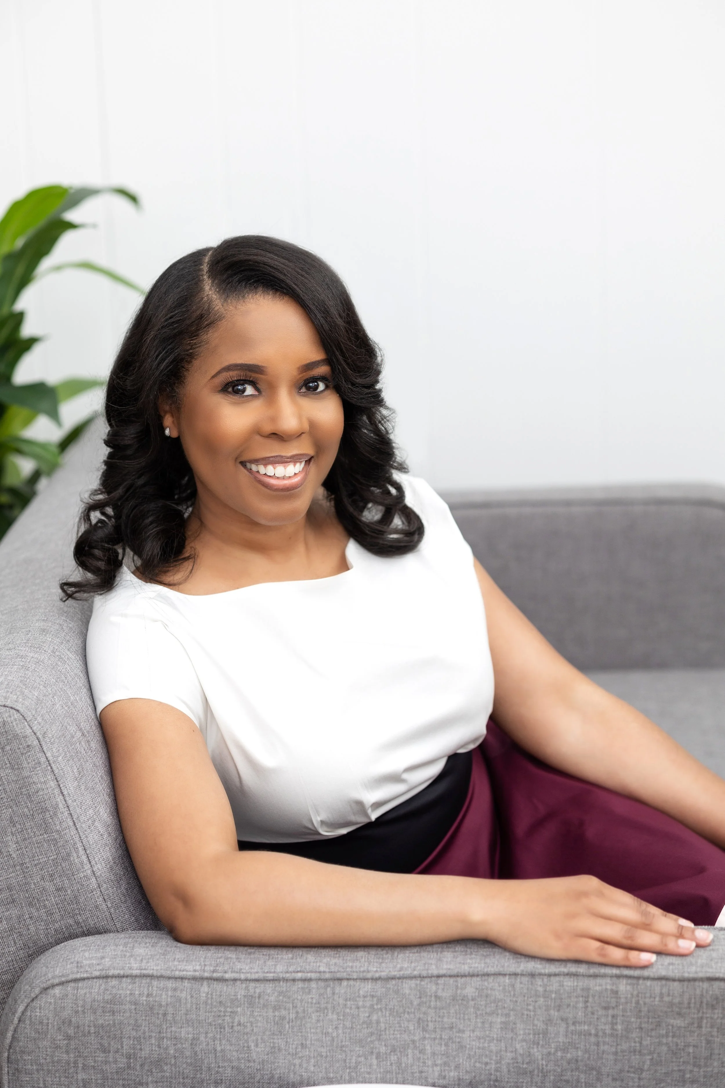 Stefani Hudson, MD, Life coach with dark, curly hair wearing a white top and maroon skirt, sitting on a gray couch in a bright room with a green plant in the background.