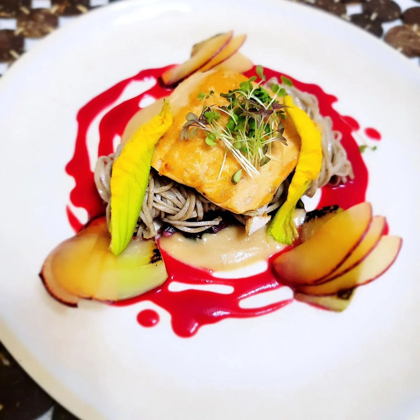 Plate of cooked fish with pasta, garnished with microgreens, peach slices, roasted apple slices, and red sauce artfully drizzled on white plate.