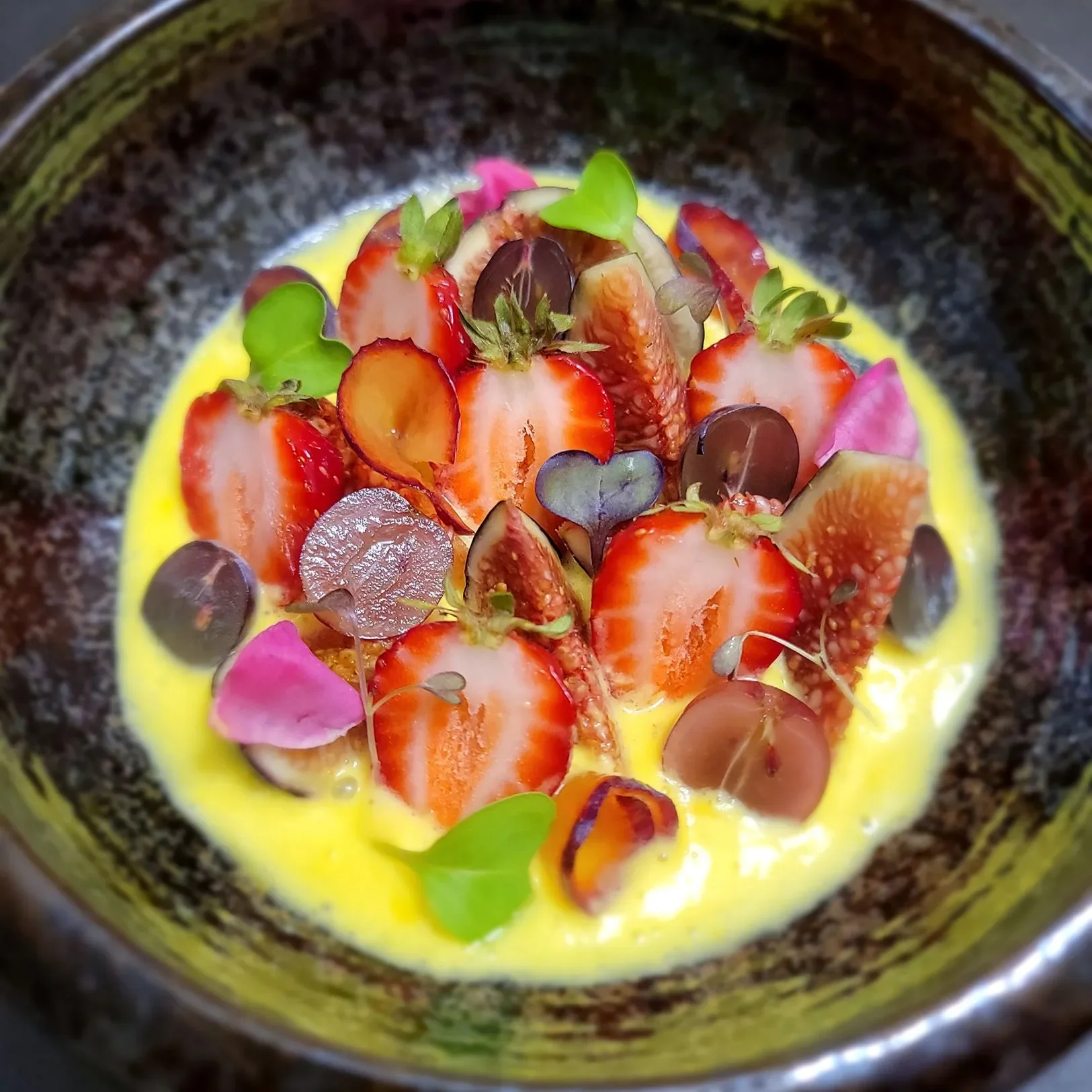 Close-up of a dessert plate with sliced strawberries, edible flowers, and small purple leaves in a yellow sauce or cream, in a dark bowl.
