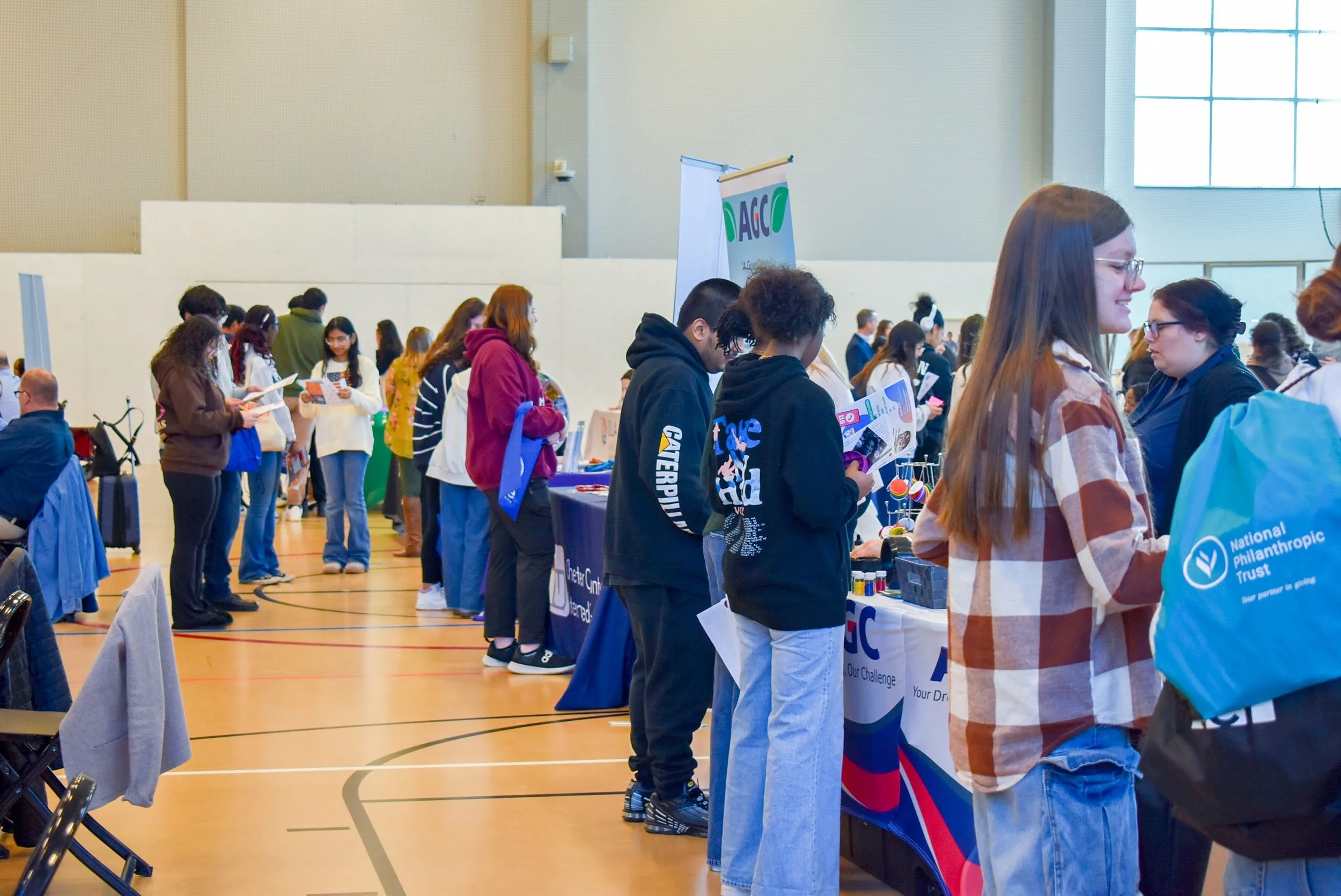 People standing in line at a community fair or career expo in a gymnasium, browsing booths with informational materials and promotional items.