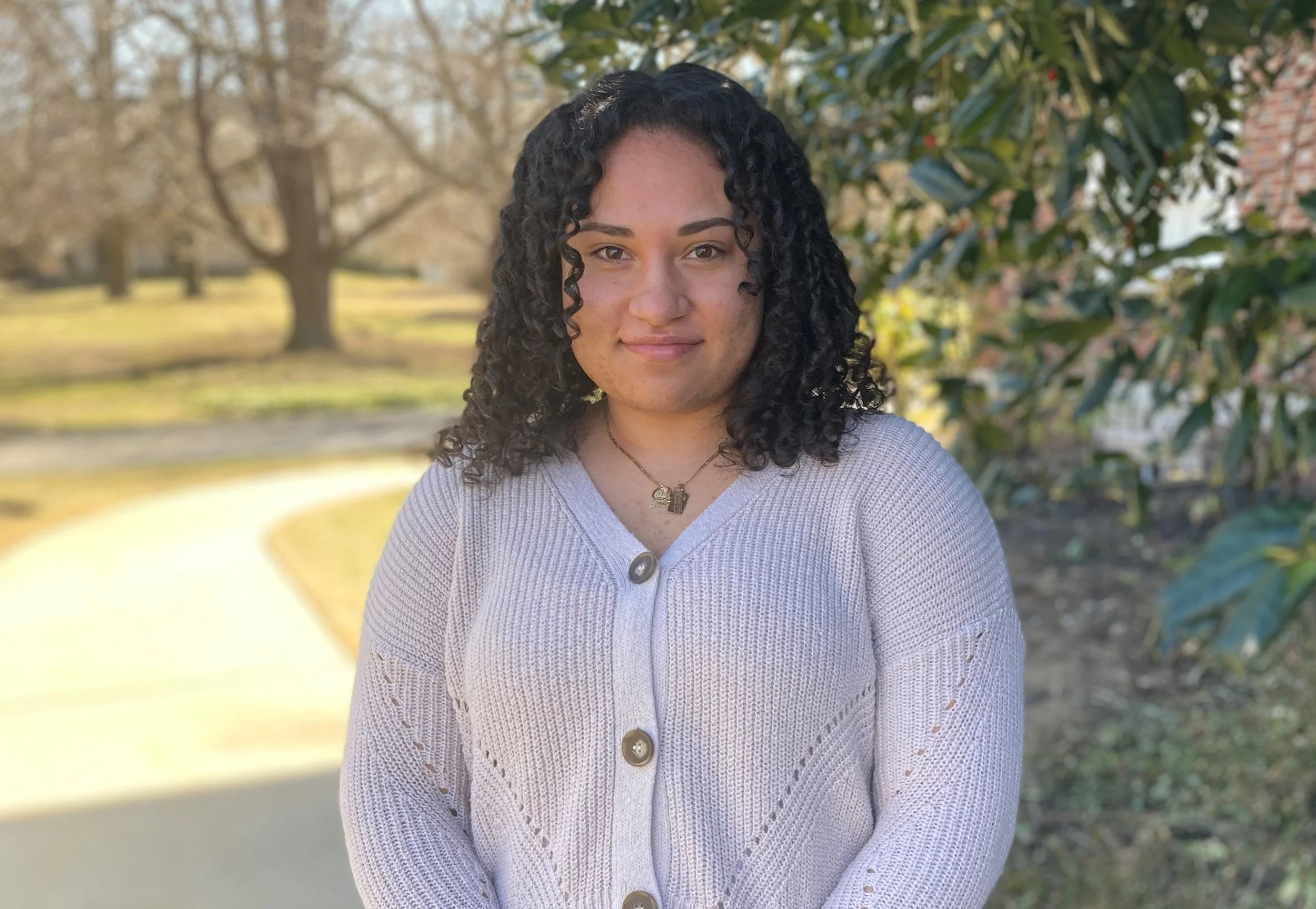 A woman with curly hair standing outdoors on a sunny day, wearing a light-colored cardigan with large buttons and a necklace, with trees and bushes in the background.