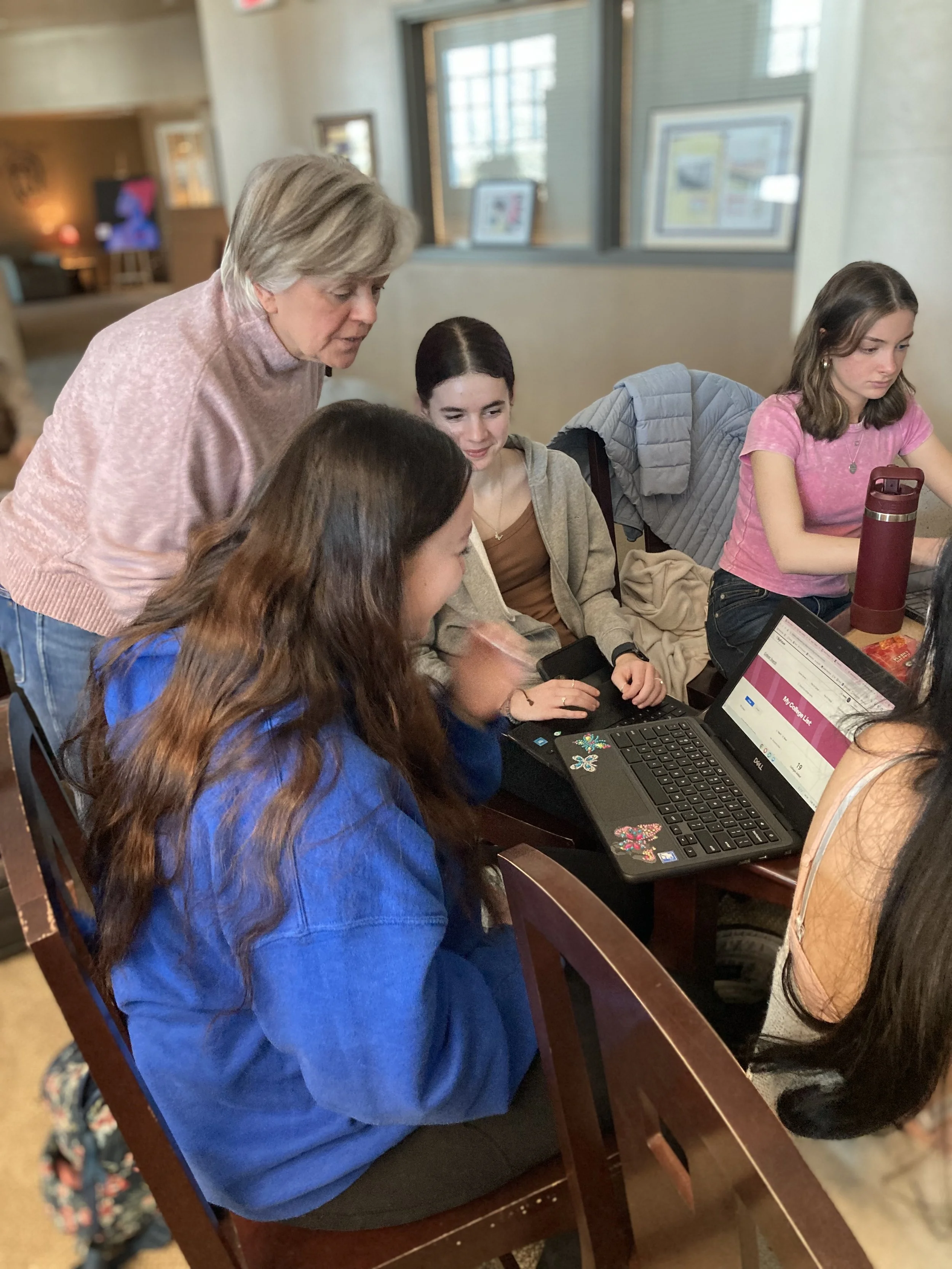 A group of young women and an older woman gathered around a table looking at a laptop in a cozy indoor space.
