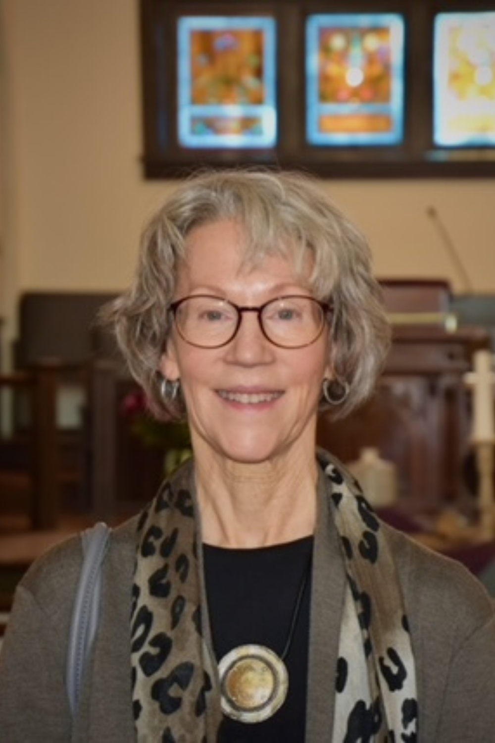Smiling elderly woman with glasses, short curly gray hair, wearing a black top, a gray cardigan, a leopard print scarf, and a large round pendant necklace, standing inside a church with stained glass windows in the background.