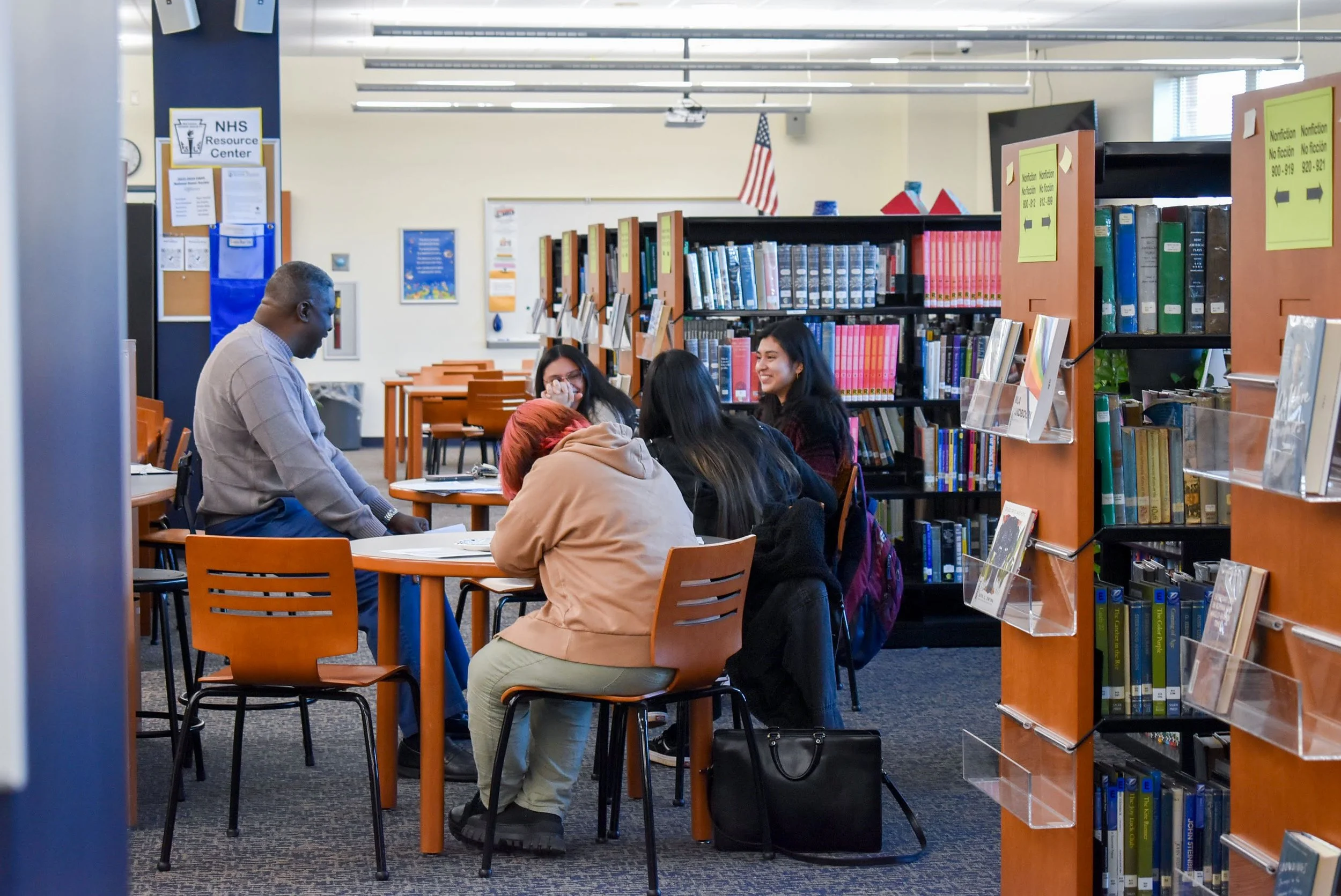 A group of five diverse people sitting and standing around a table in a library, engaging in conversation, with bookshelves in the background.